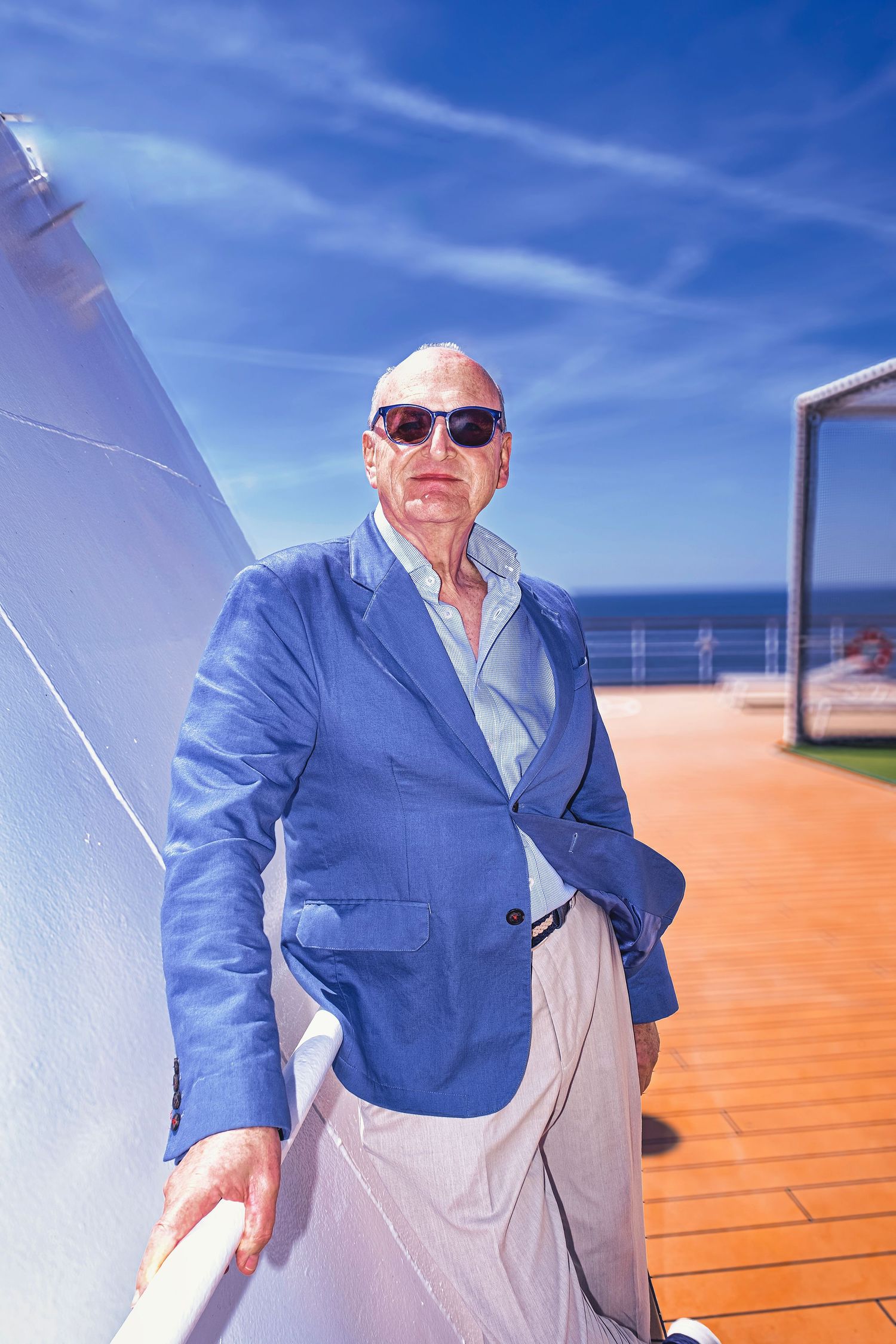Elegant bald senior man in blue blazer and sunglasses posing on cruise ship deck with ocean view.