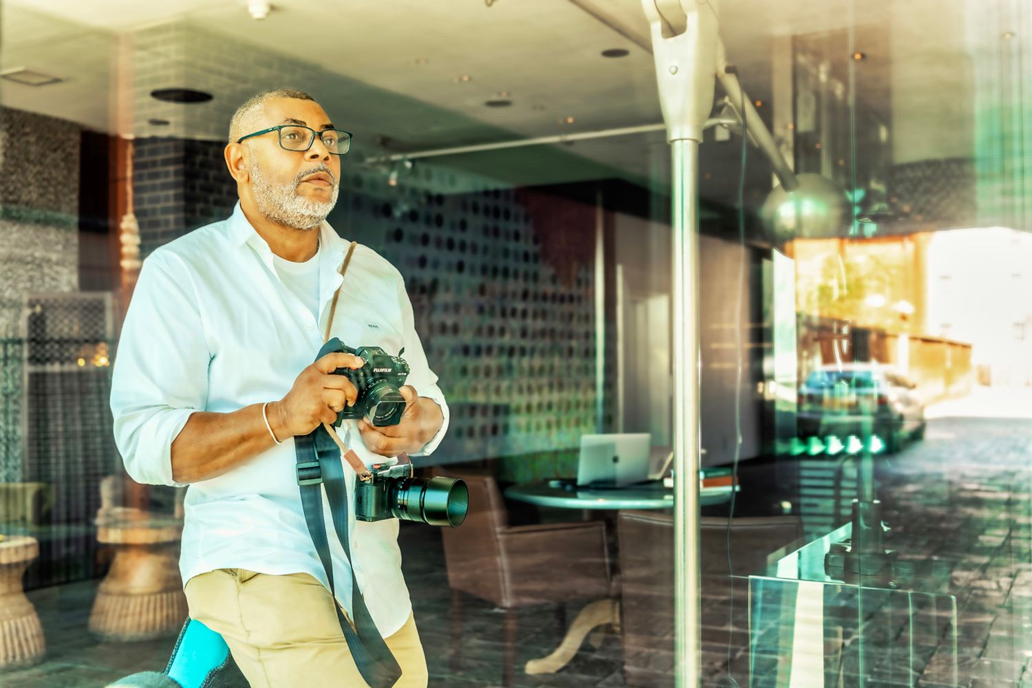 Older man with glasses holding a camera indoors, capturing photos in a modern lobby setting with natural light.