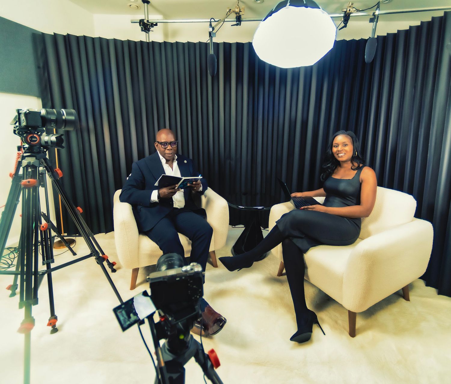 Two professionals sit in white chairs during a TV studio interview, with cameras and studio lighting visible.
