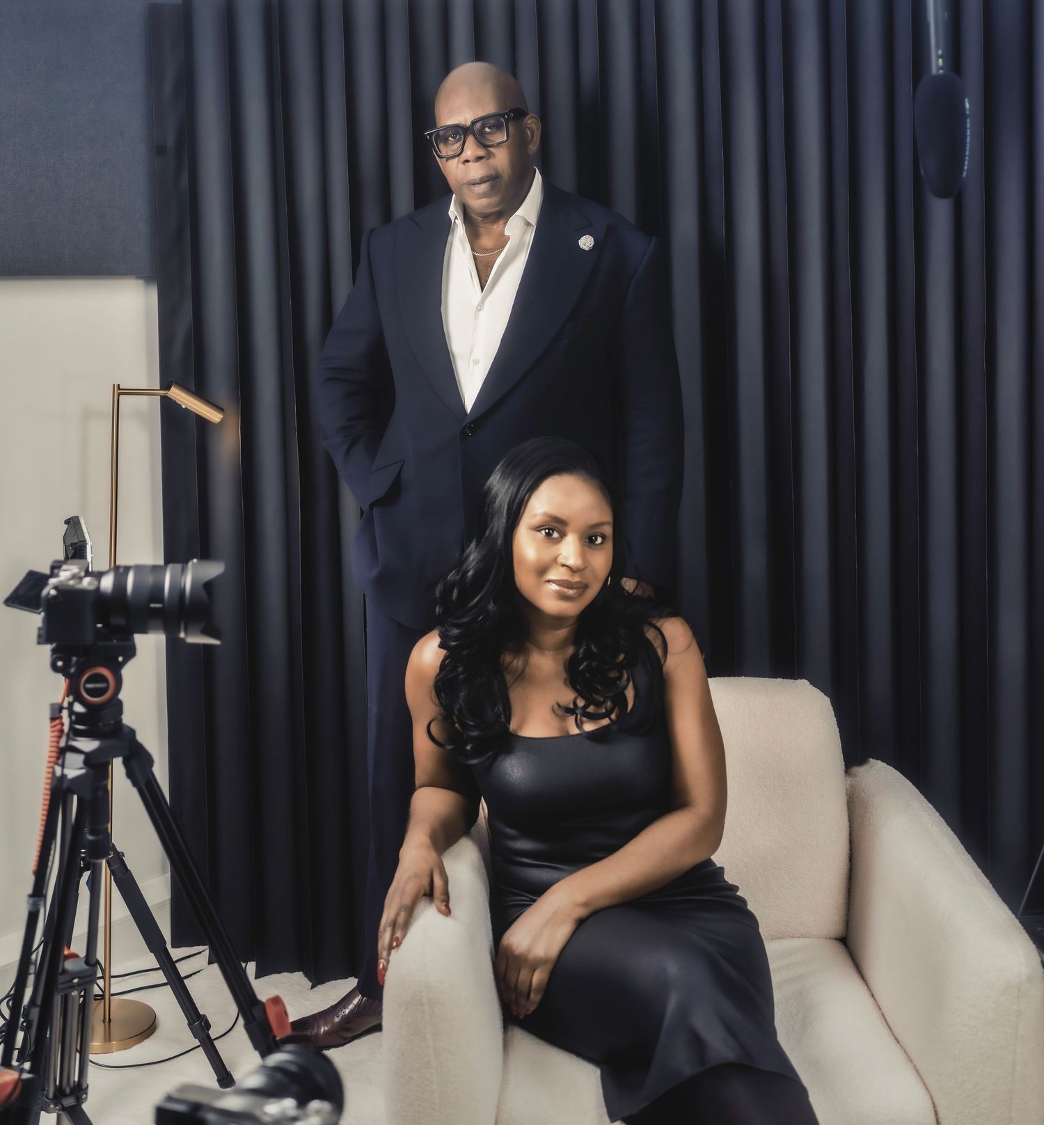 A man in a navy suit stands behind a woman in a black dress seated in a white chair in a studio setting.