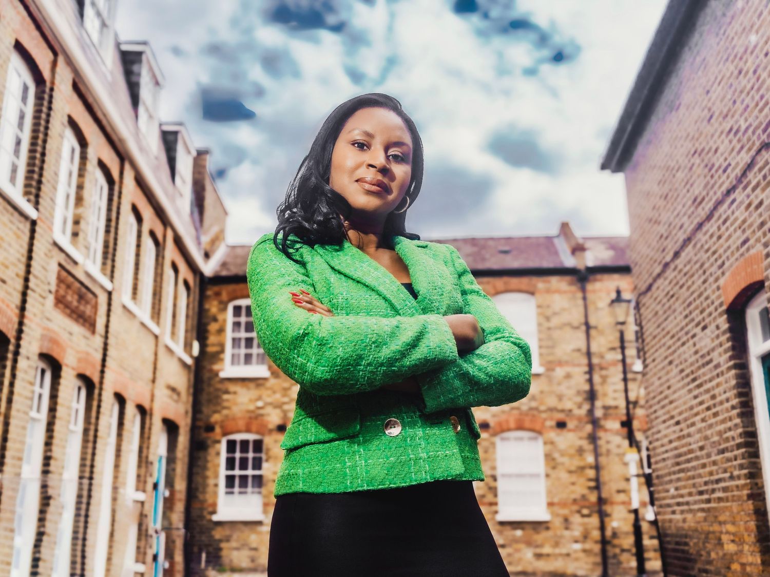 Professional woman in green blazer standing confidently with arms crossed in a brick alleyway.