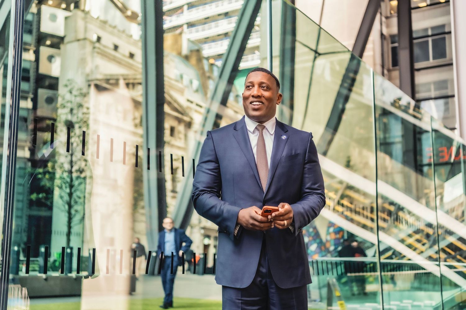 Professional businessman in navy suit smiling outdoors near modern glass building in urban city setting.