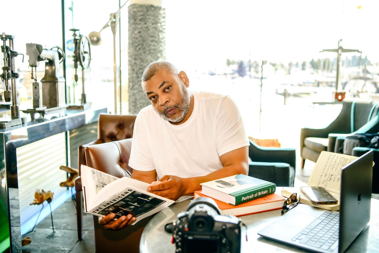 Man in white t-shirt sitting at desk with camera, laptop, and books in a modern office setting.