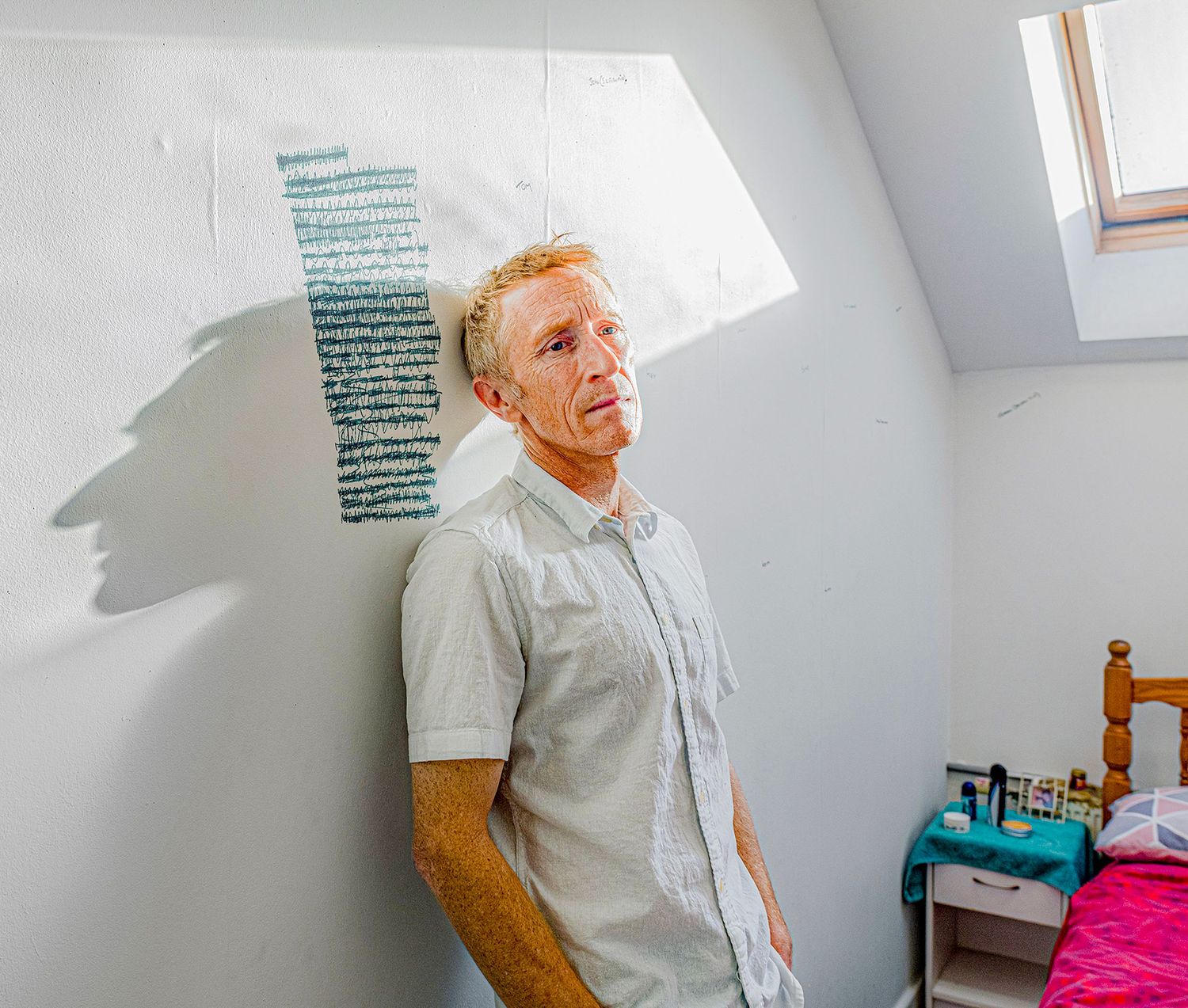 Man with blonde hair leaning against a white wall in a bedroom with a skylight and pink bed.