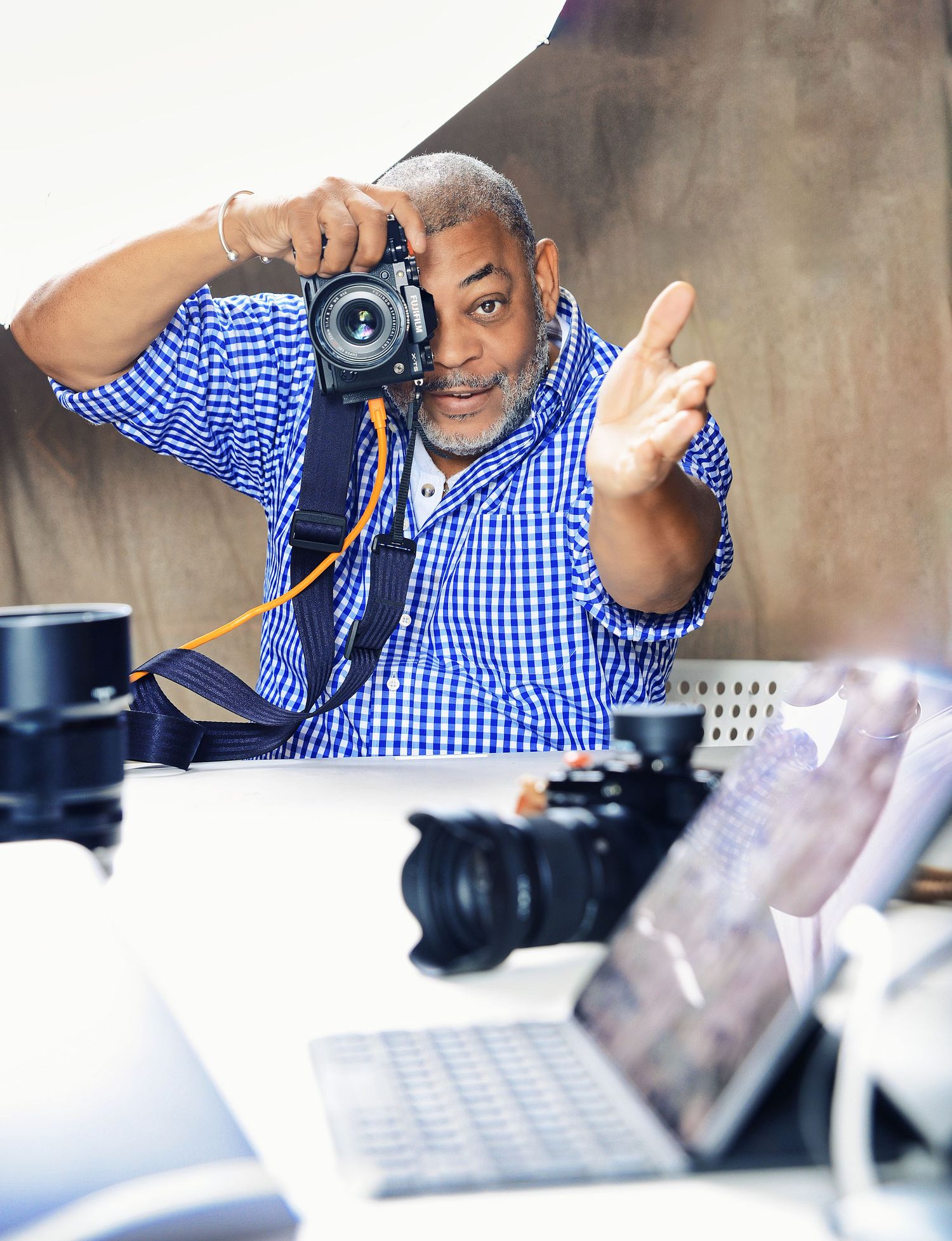 Photographer in blue gingham shirt captures shot with DSLR camera, surrounded by lenses and laptop in studio.