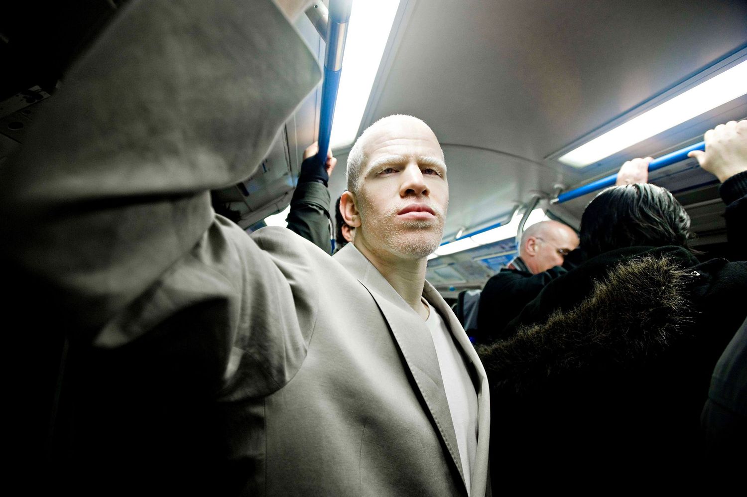 Man with albinism standing in a crowded subway train, wearing a gray jacket, surrounded by other passengers.