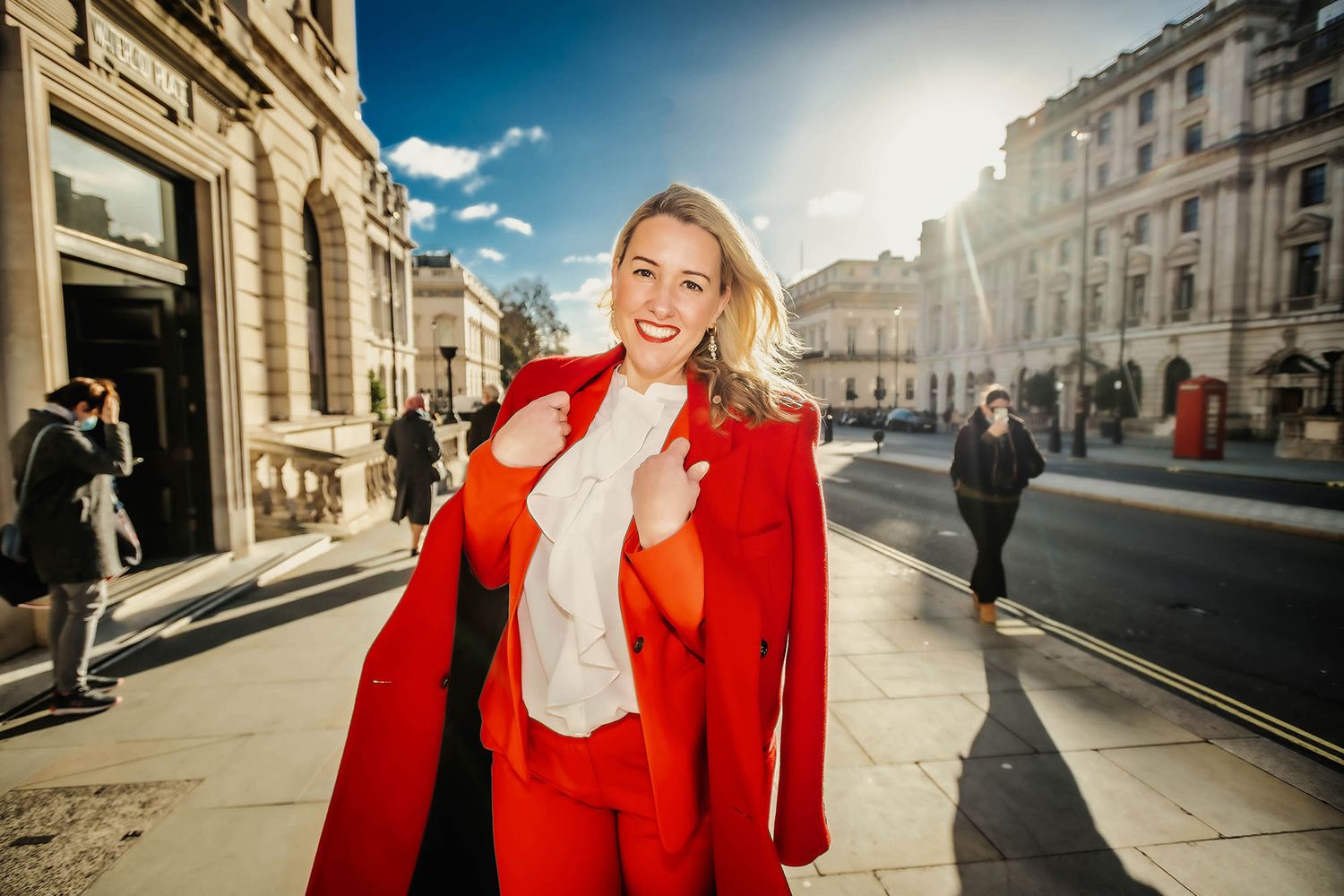 Woman in red suit walking confidently on a sunny London street, with classic architecture and a red phone box in background.