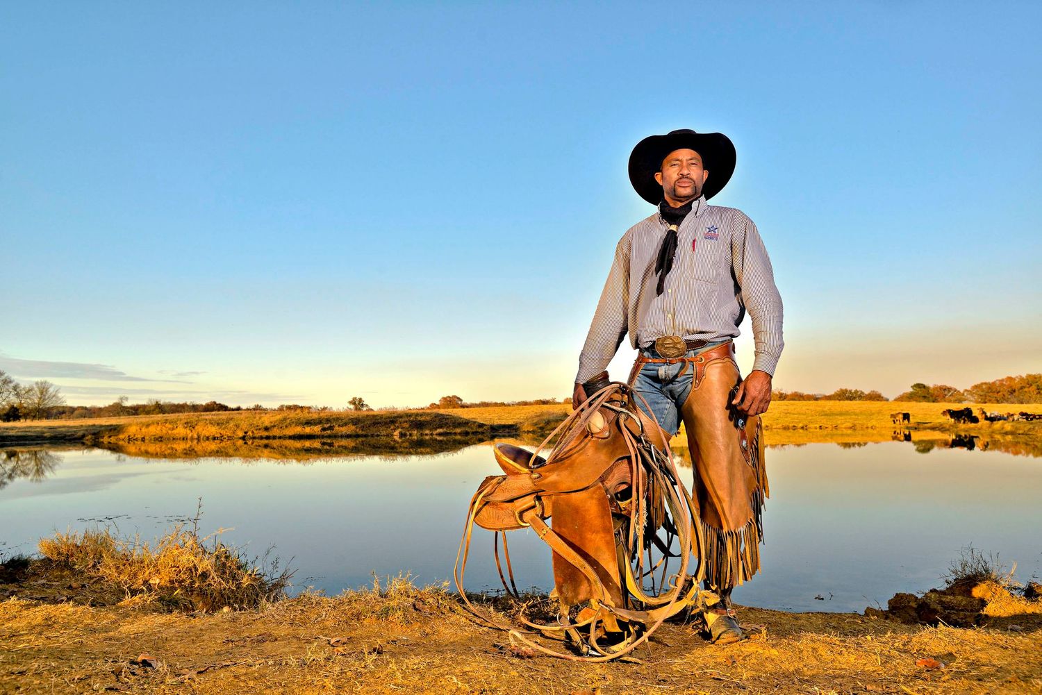 Cowboy in hat and chaps stands with saddled horse near a peaceful pond at golden hour on ranch land.
