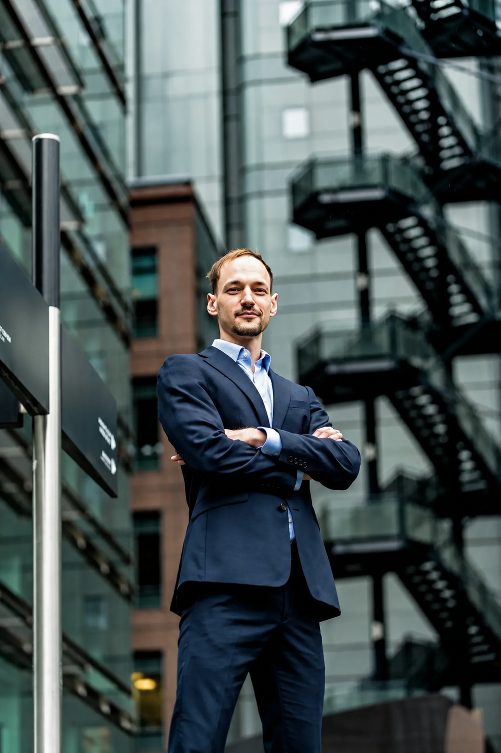 Professional businessman in navy suit standing confidently with arms crossed outside modern glass office building.