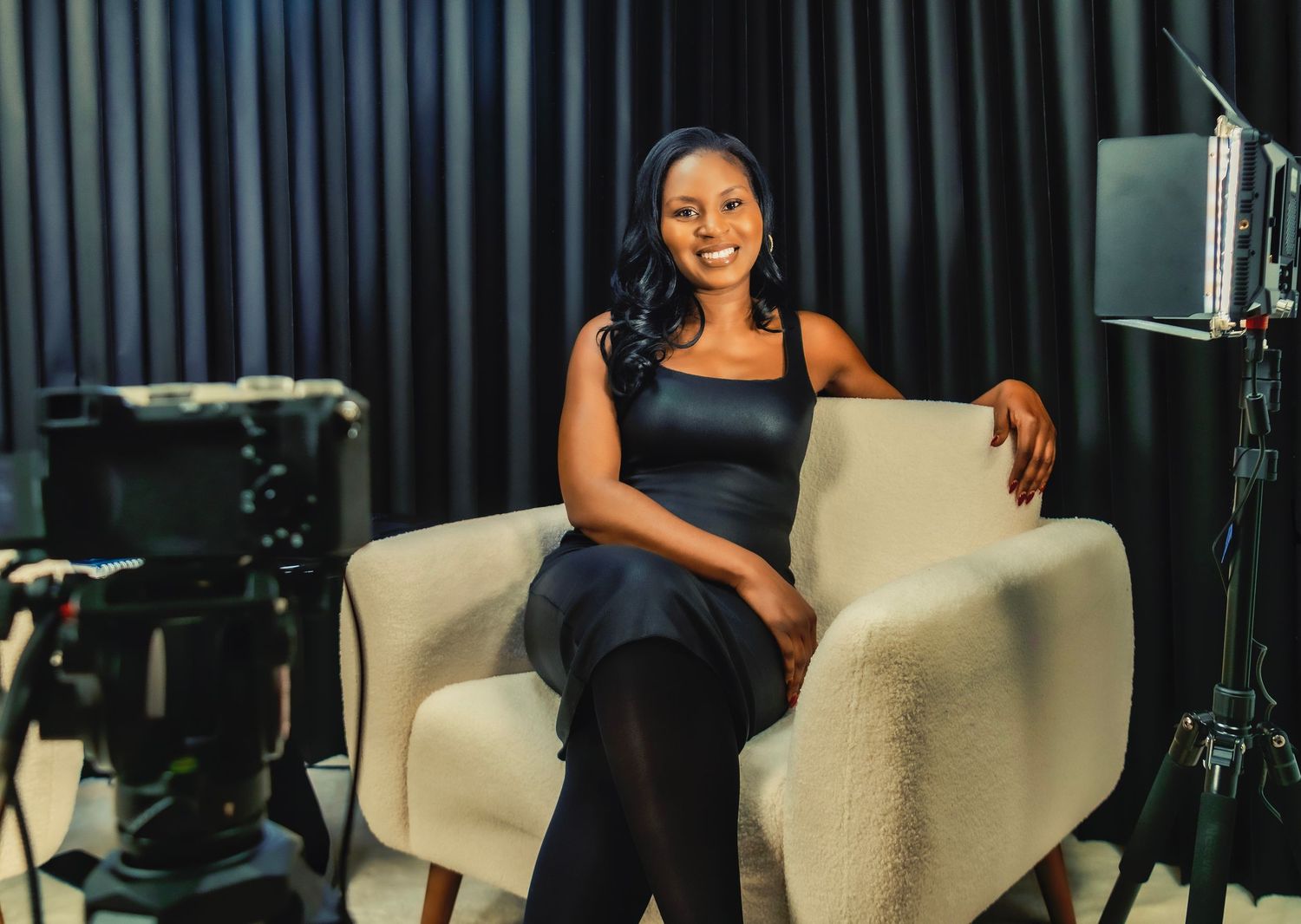 Woman in black dress smiling confidently in a professional TV studio setting with camera equipment.