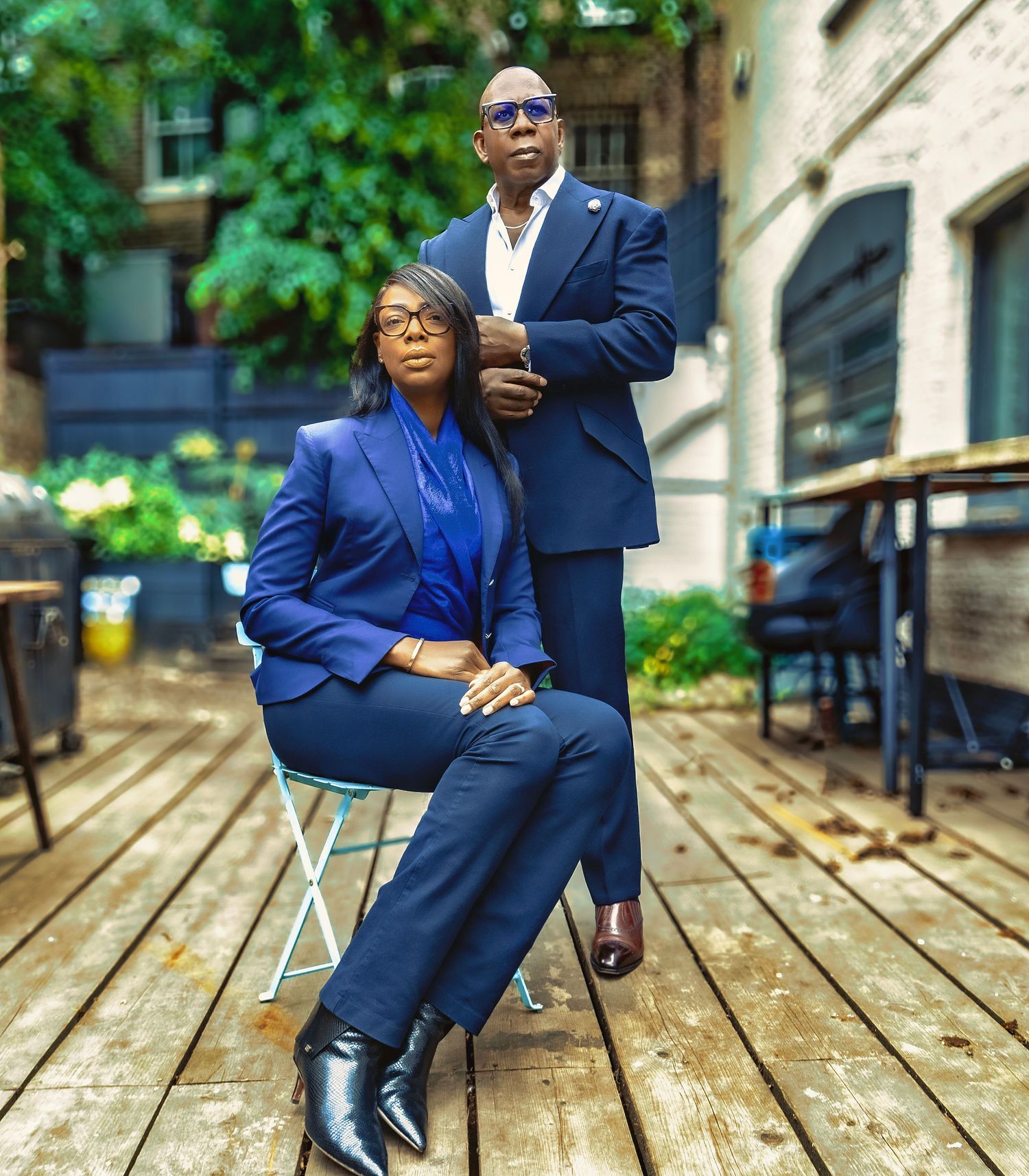 Two professionals in blue suits pose on an outdoor wooden deck, one seated and one standing, with green foliage backdrop.