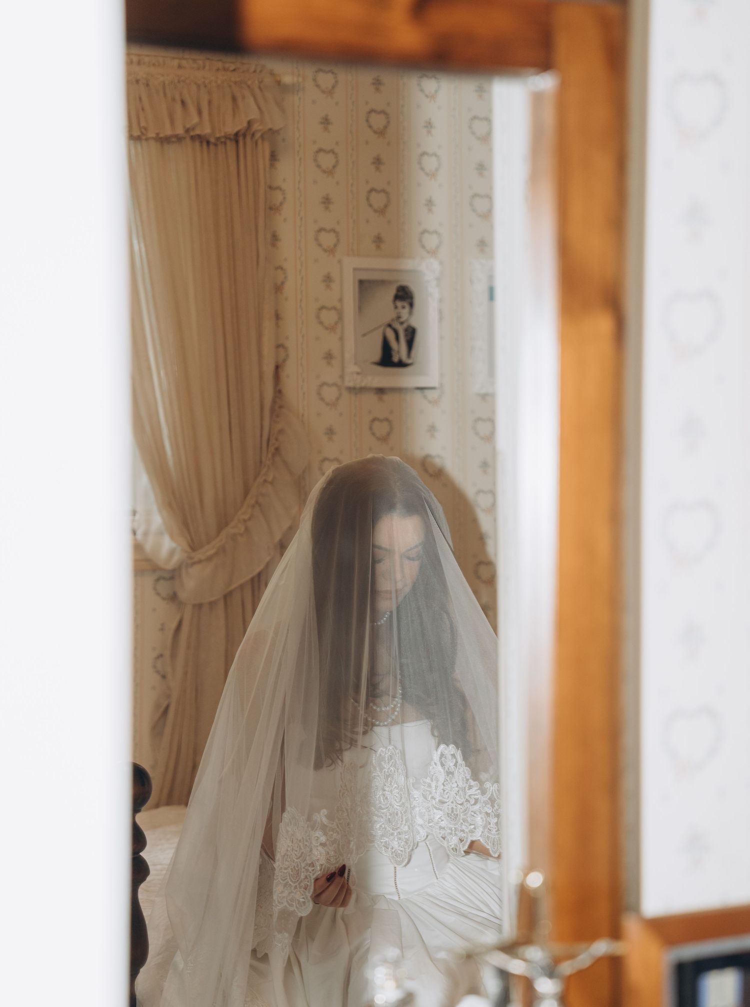 A wedding dress with delicate lace details is displayed in a cream colored room with elegant curtains.
