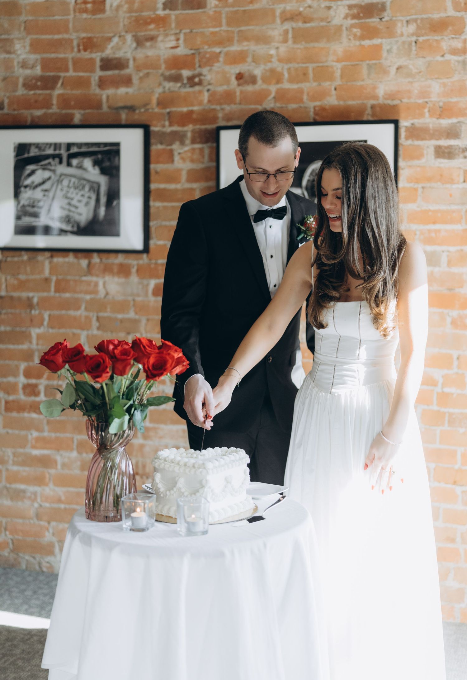 A bridal party wearing white and black formal attire performs wedding candle lighting ceremony at brick wall venue.