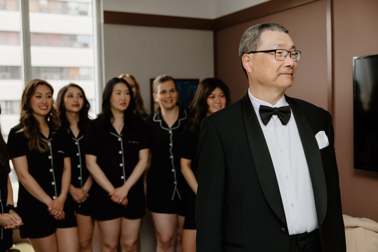 A formal man in tuxedo and bow tie stands in front of a group wearing matching black outfits.