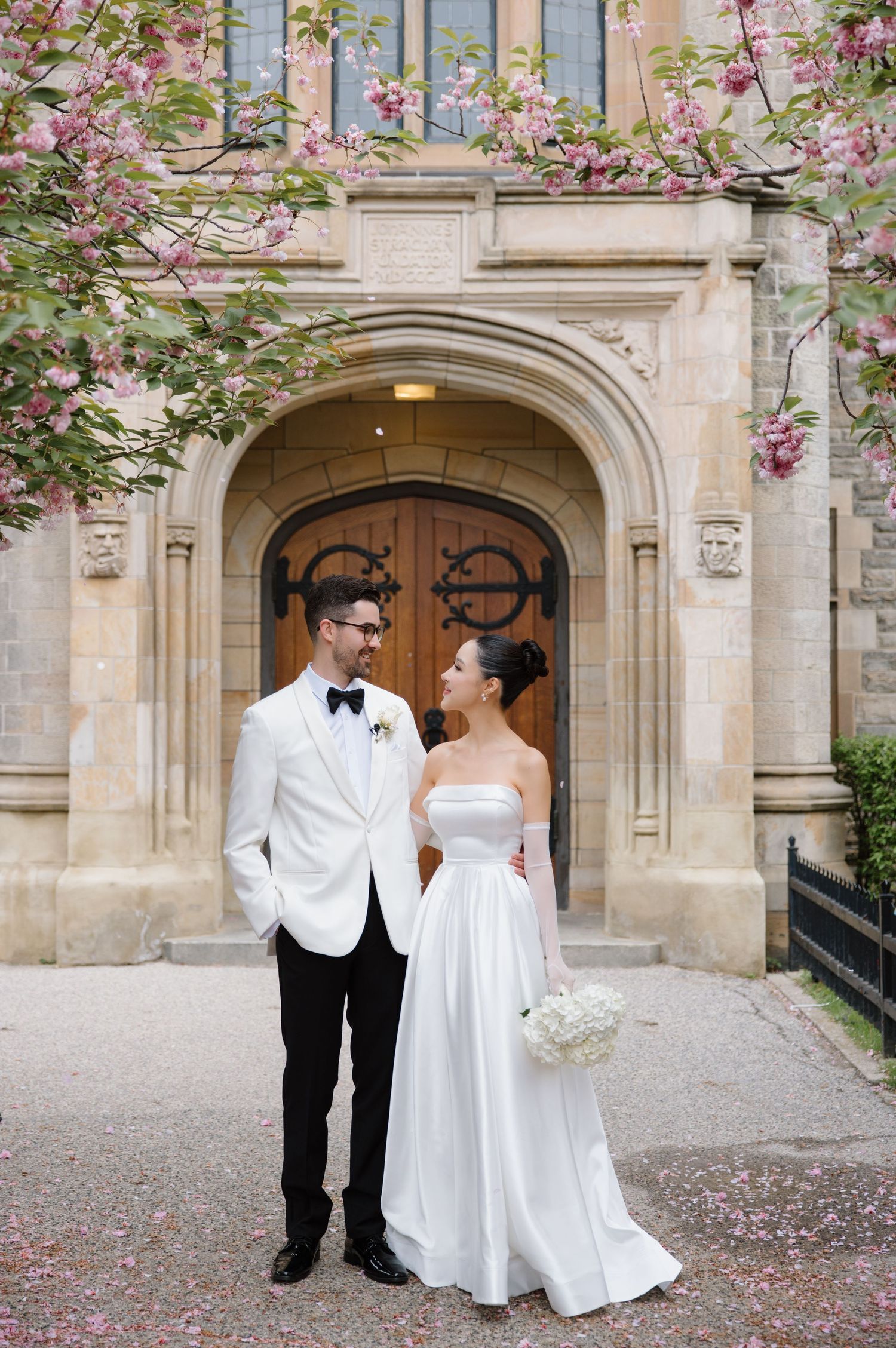A wedding couple stands in front of an arched stone doorway with pink cherry blossoms blooming overhead.