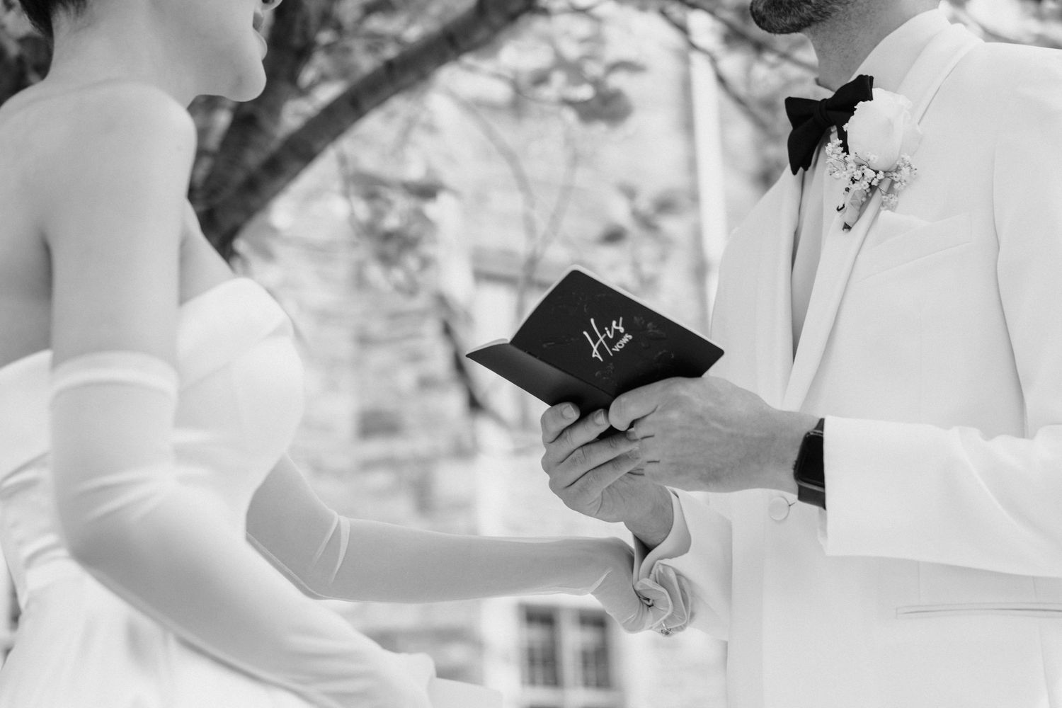 Black and white photo of wedding ceremony with officiant holding prayer book while couple exchanges vows outdoors.