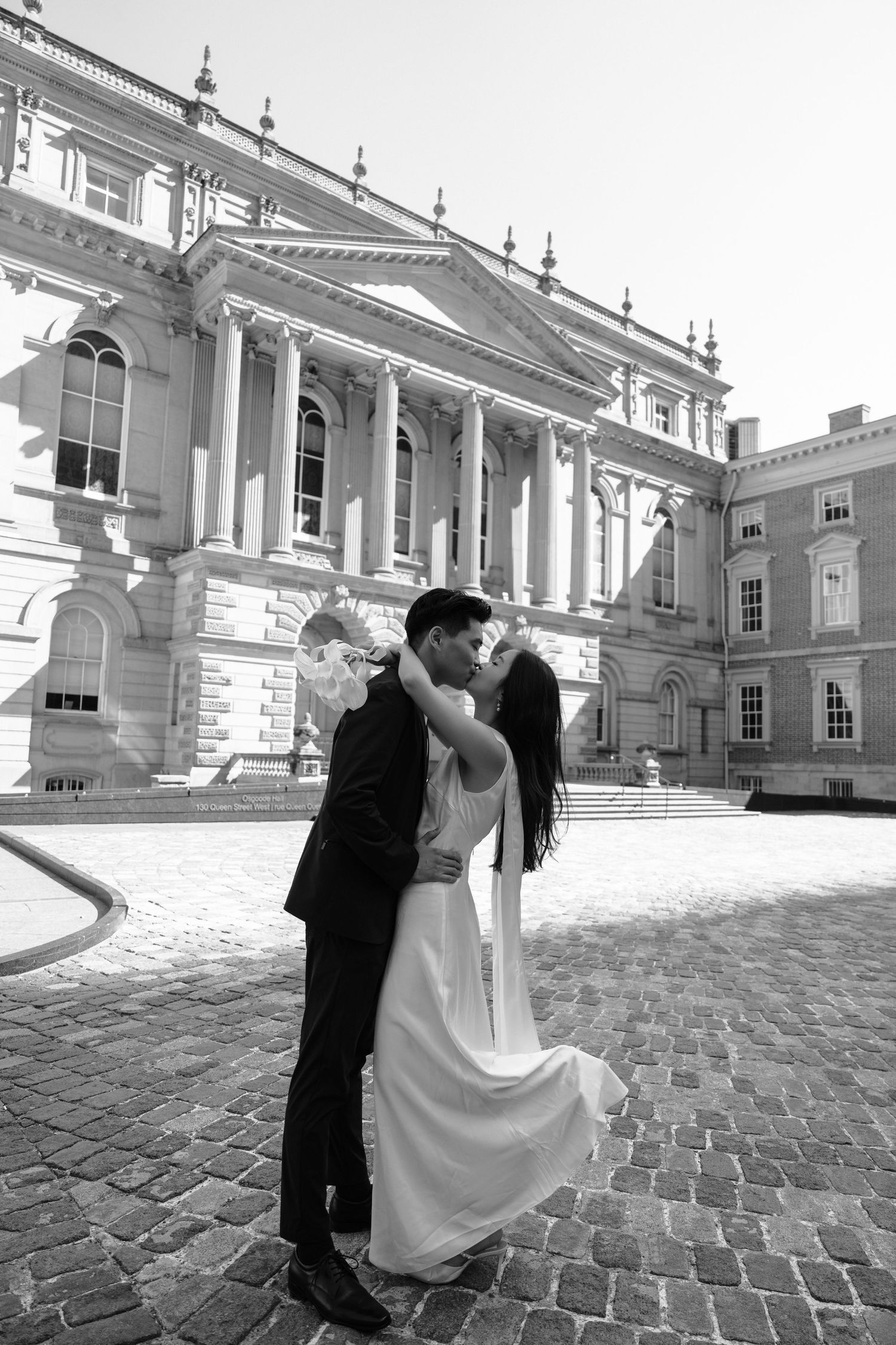 A romantic couple shares a kiss outside an elegant neoclassical building with white columns and ornate architecture.