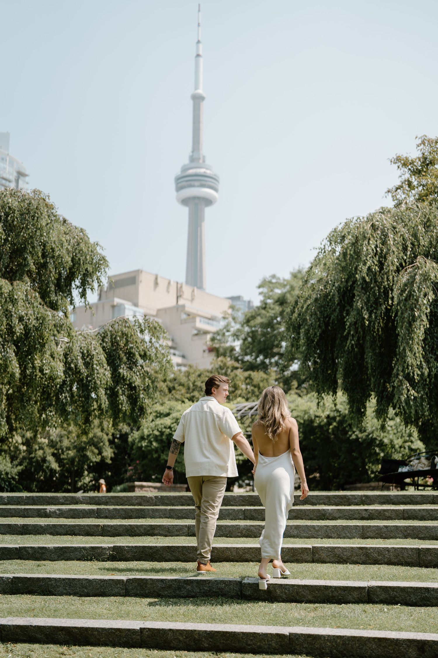 A couple walks together on green grass with Toronto's CN Tower and city skyline visible in the background.