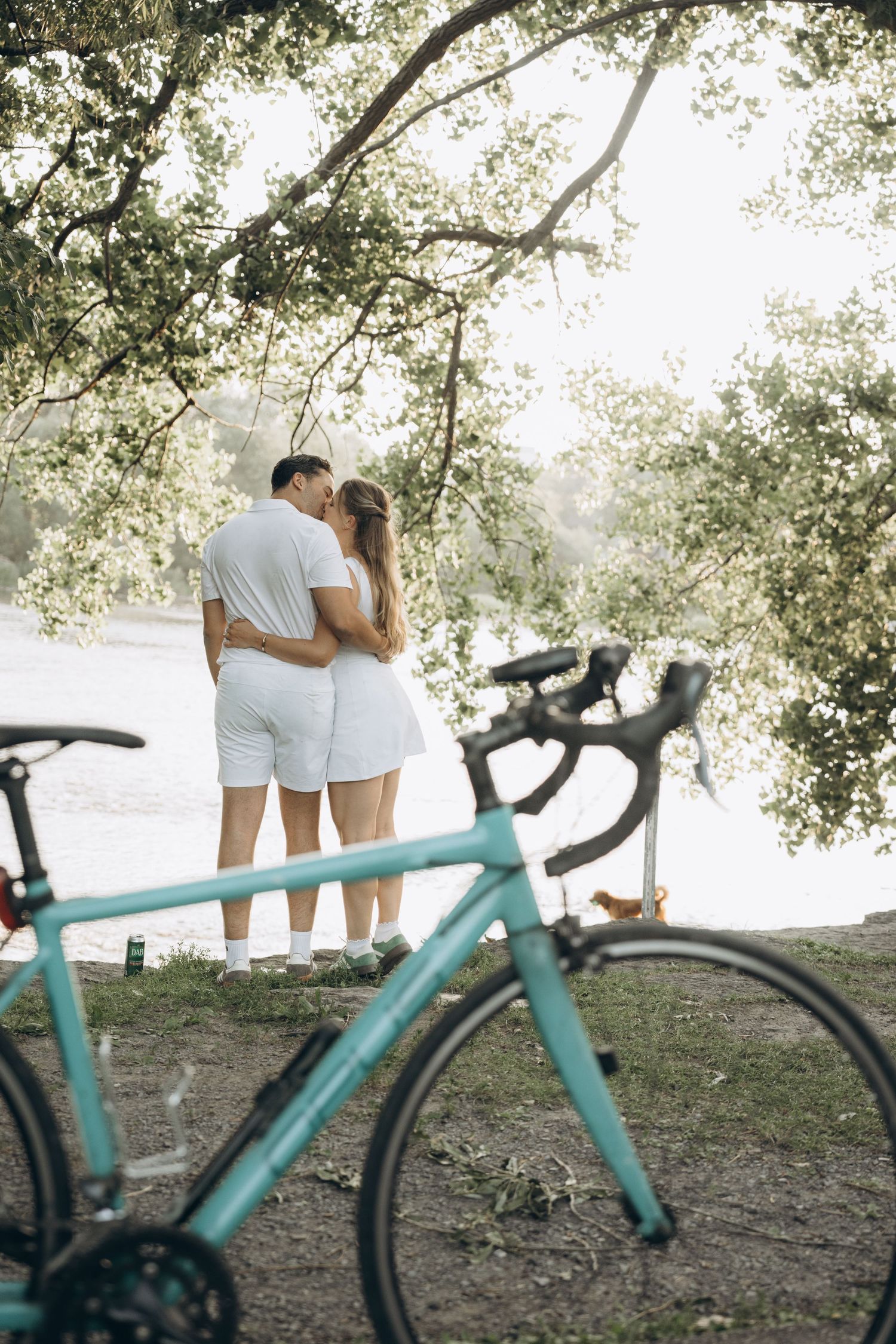 A turquoise bicycle stands in foreground while a couple in white clothing embraces beneath shady trees on a summer day.
