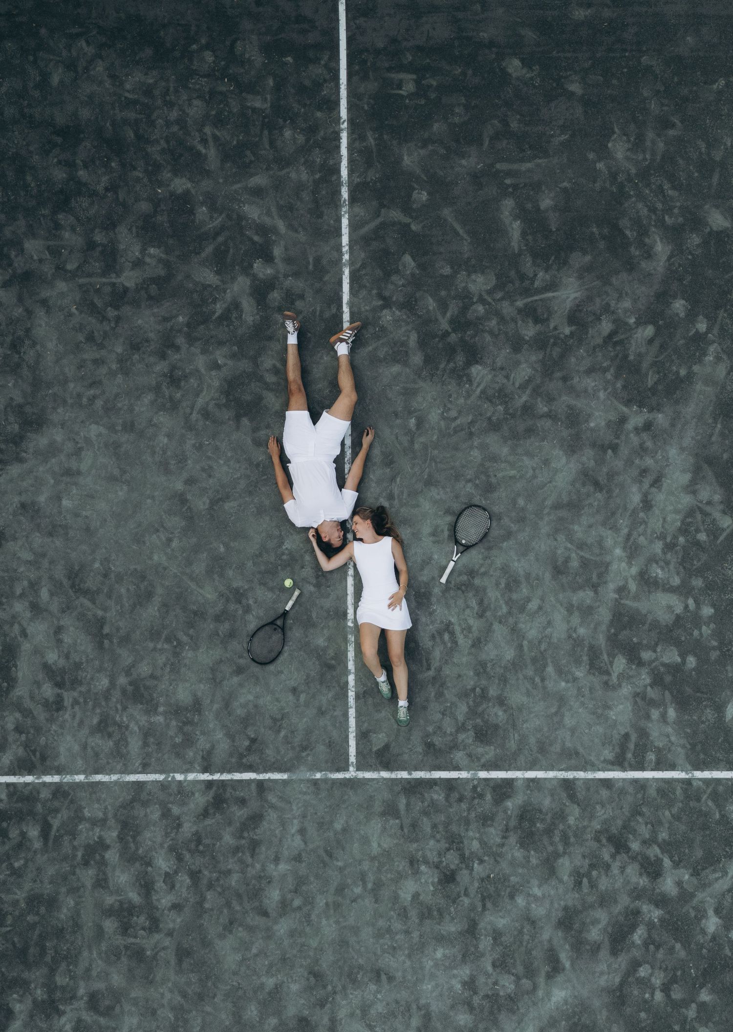 Tennis player in white attire lies defeated on a hard court after an intense match point, viewed from above.