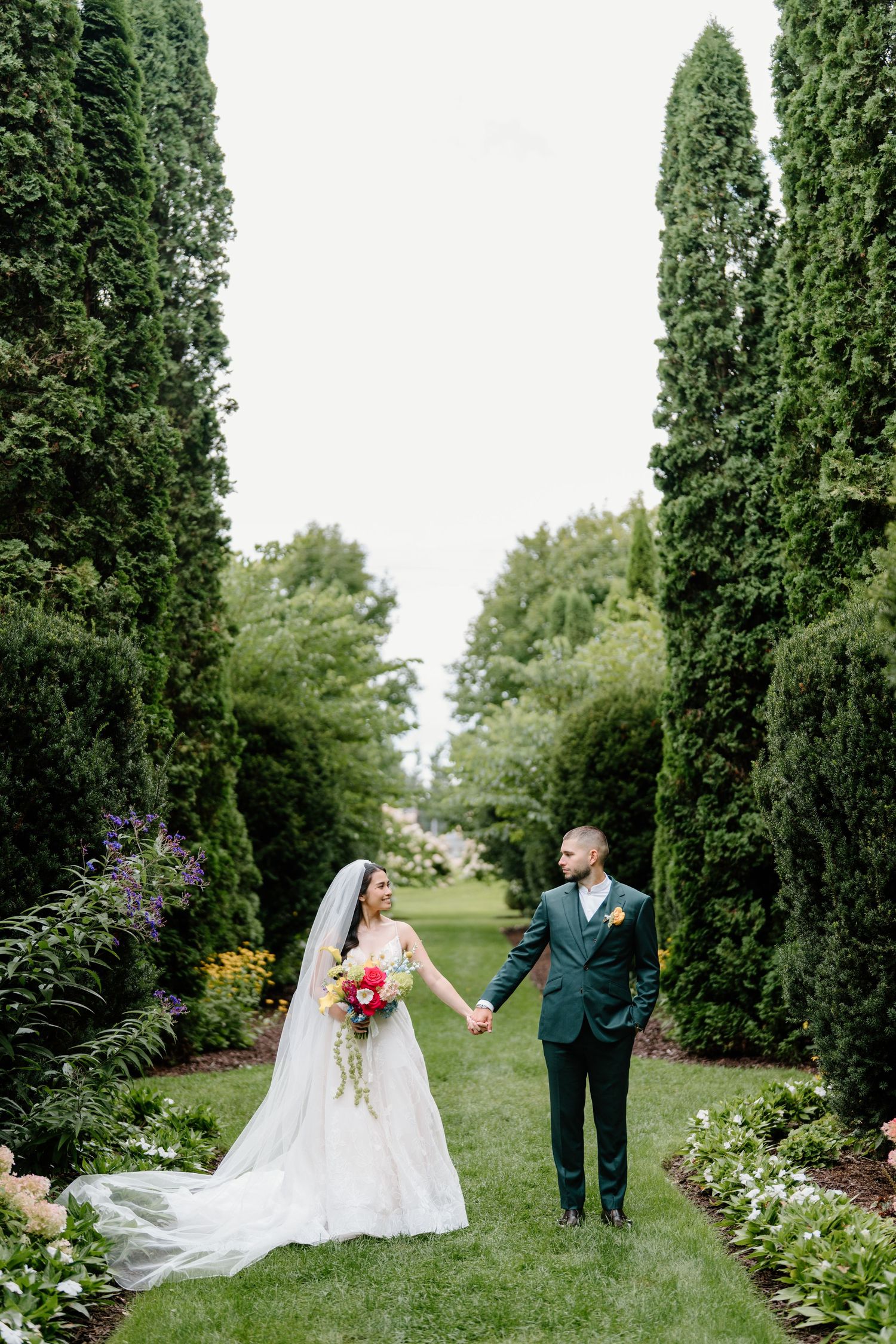 Newlyweds share romantic moment on garden path surrounded by lush greenery and cypress trees.