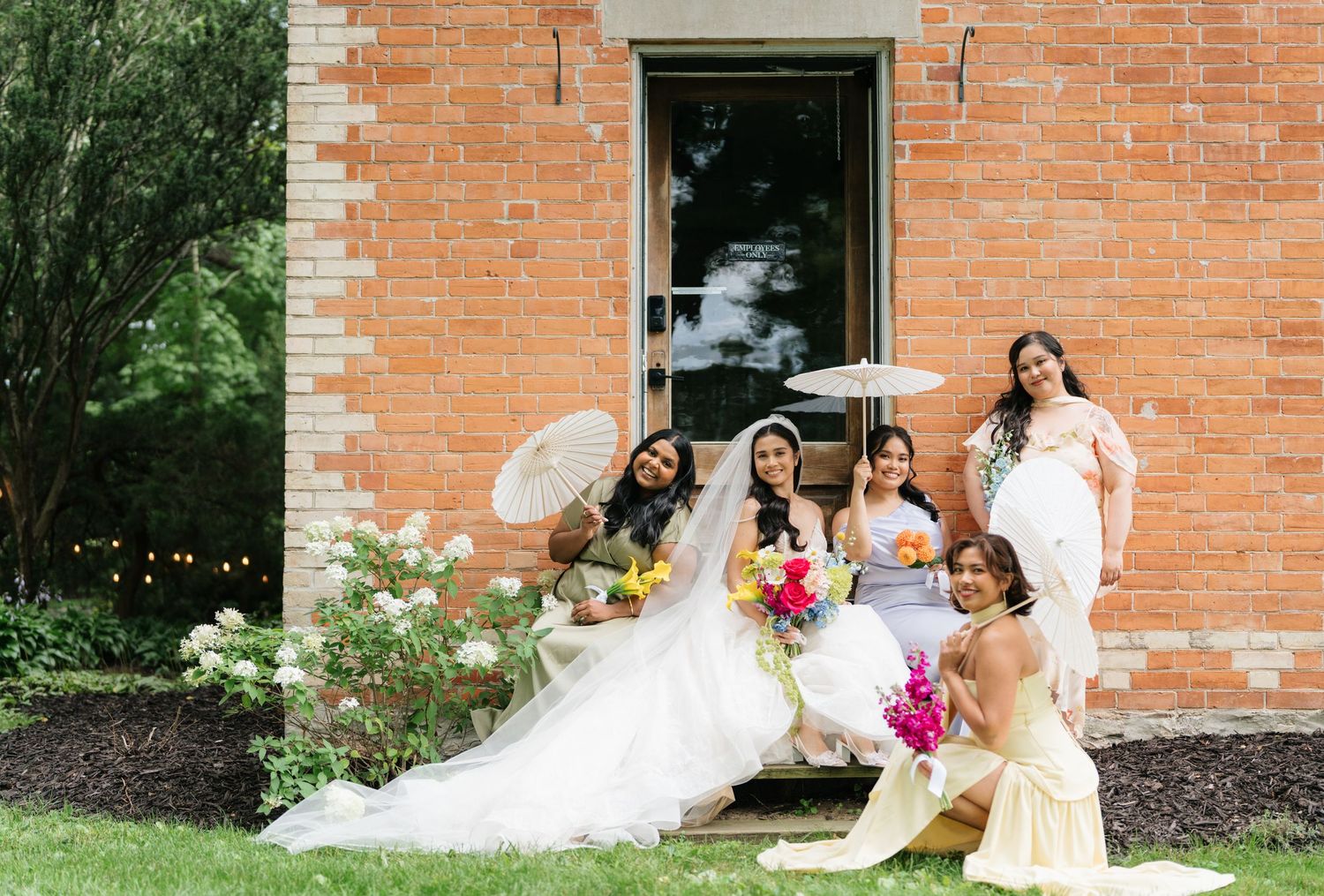 A wedding party poses in front of a brick building with white trim during an outdoor summer celebration.