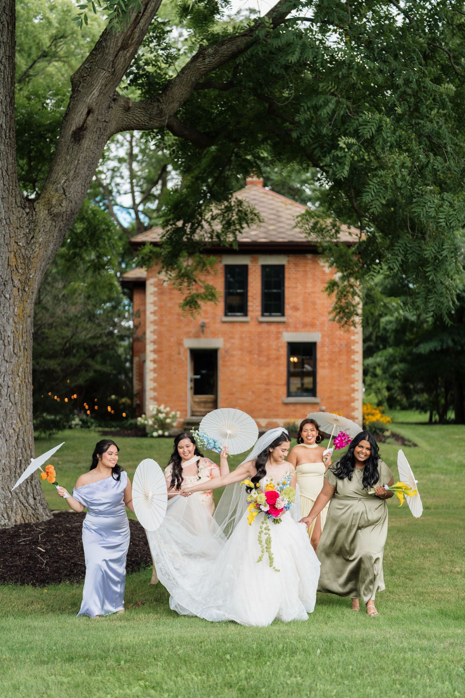A group in formal attire playfully poses on a grassy lawn with trees and a brick building in the background.