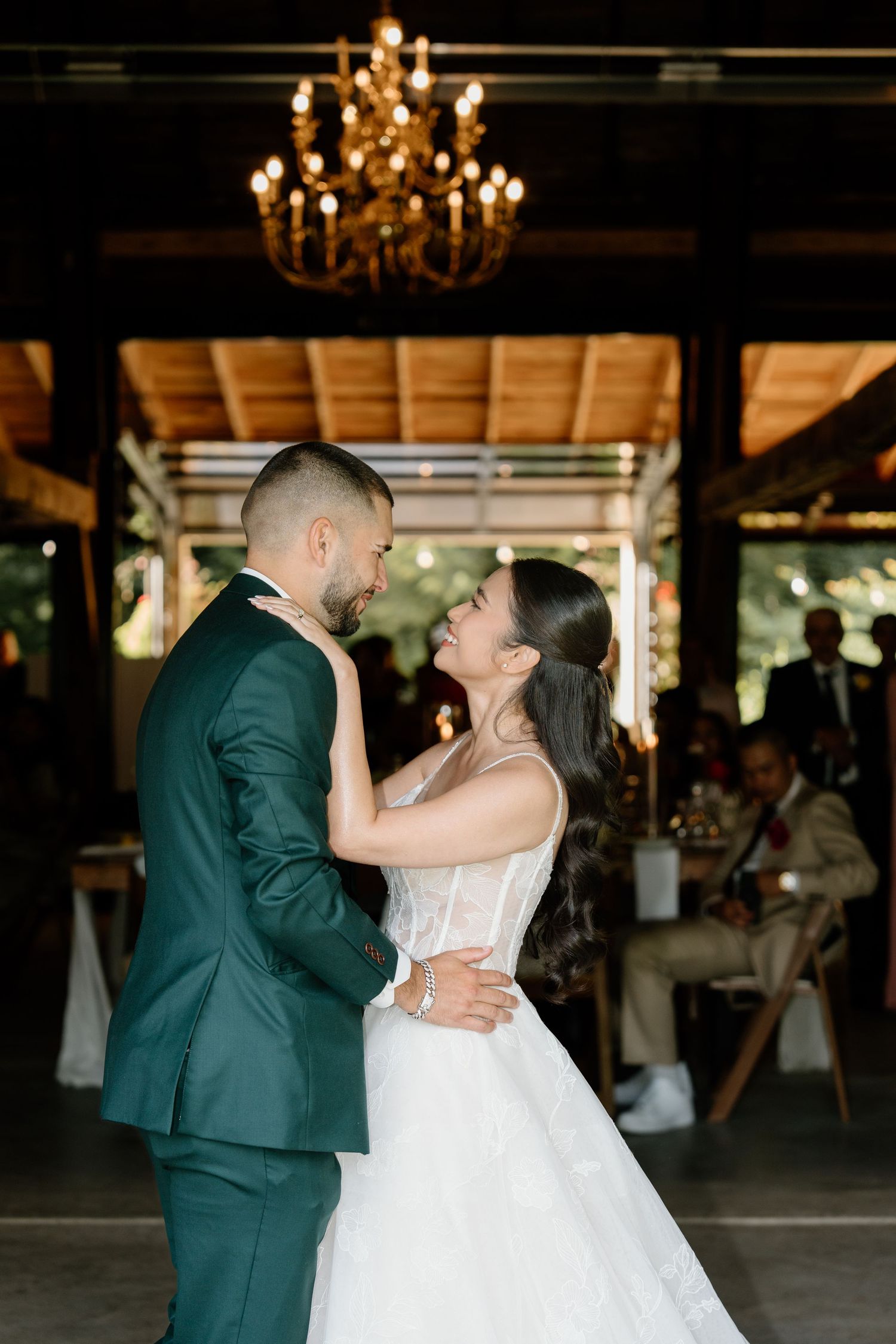 A couple shares their first dance under an elegant chandelier at a rustic barn wedding venue.
