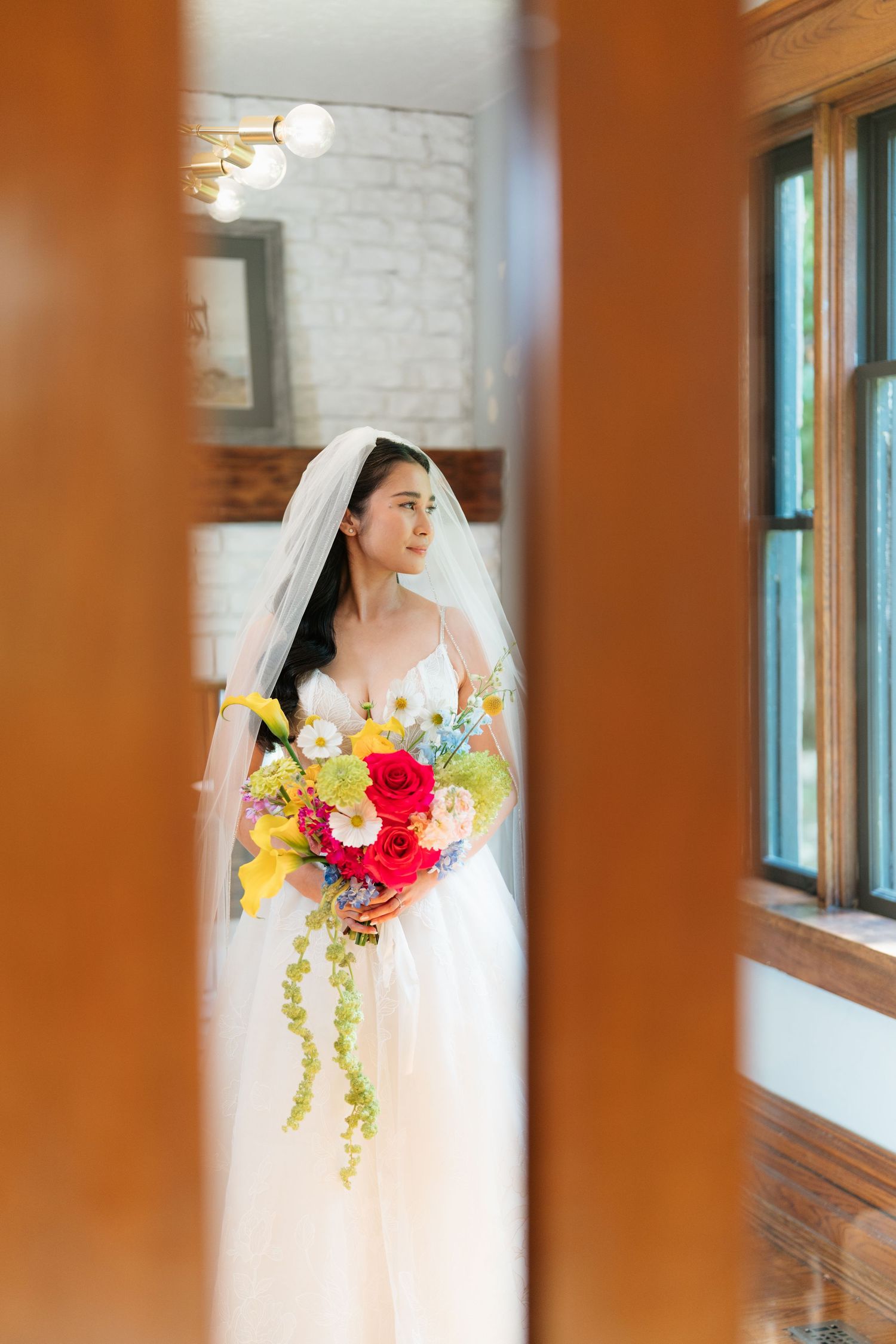 A bride wearing a traditional white veil stands in an elegant indoor space with warm lighting and wooden details.