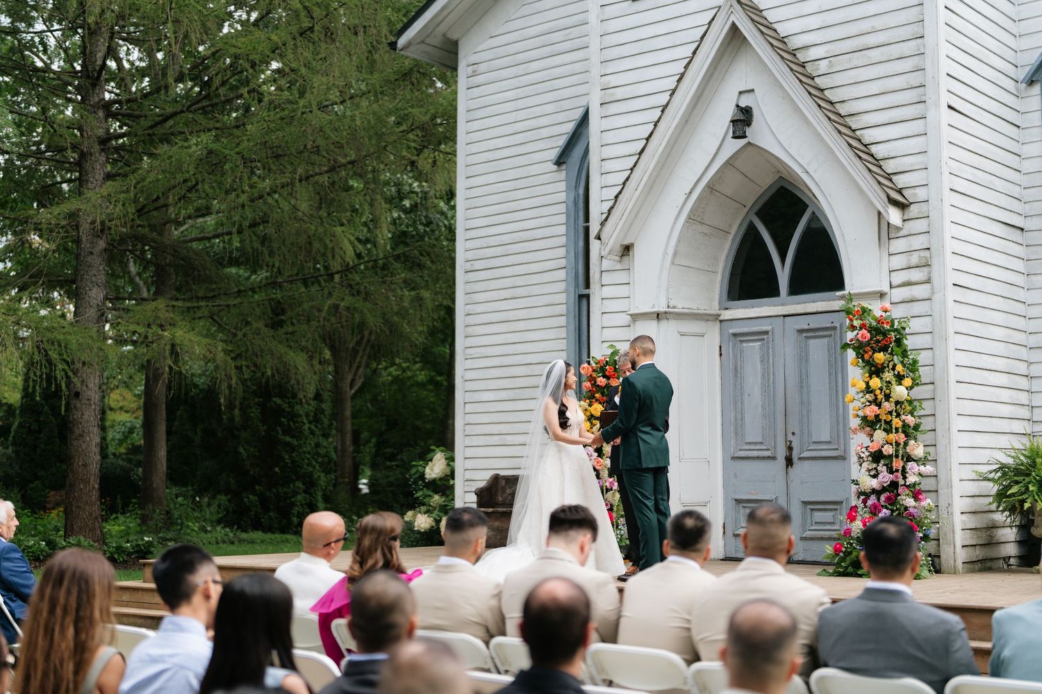 A traditional white wooden church with Gothic arched windows hosts an outdoor wedding ceremony on a sunny day.