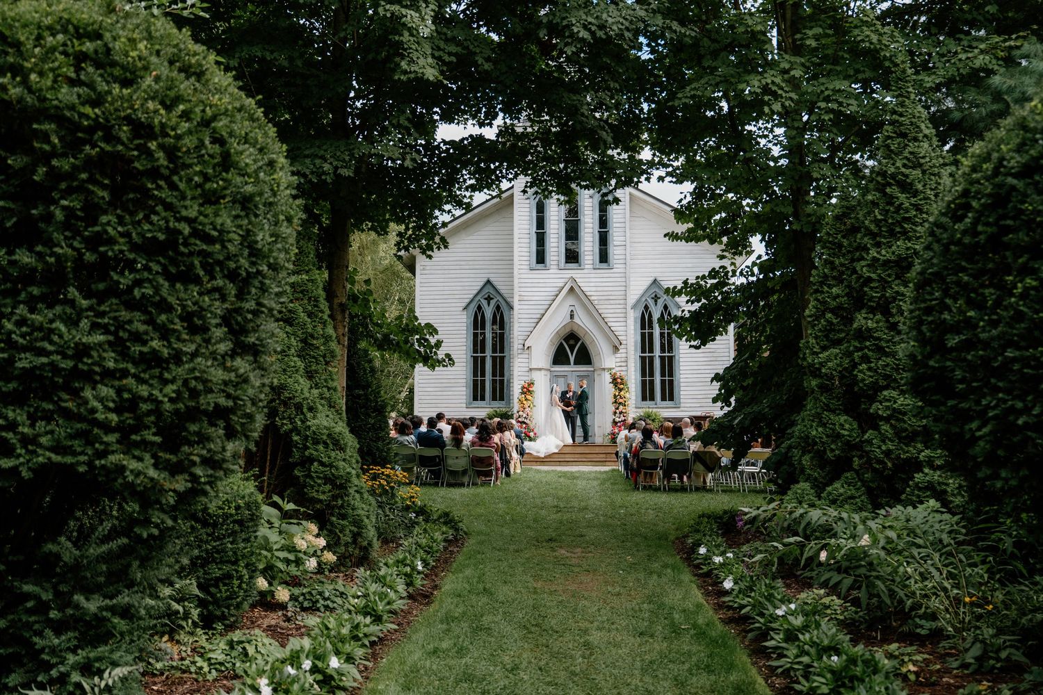 A wedding ceremony setup outdoors with white chairs arranged in rows leading to a white chapel through a garden path.