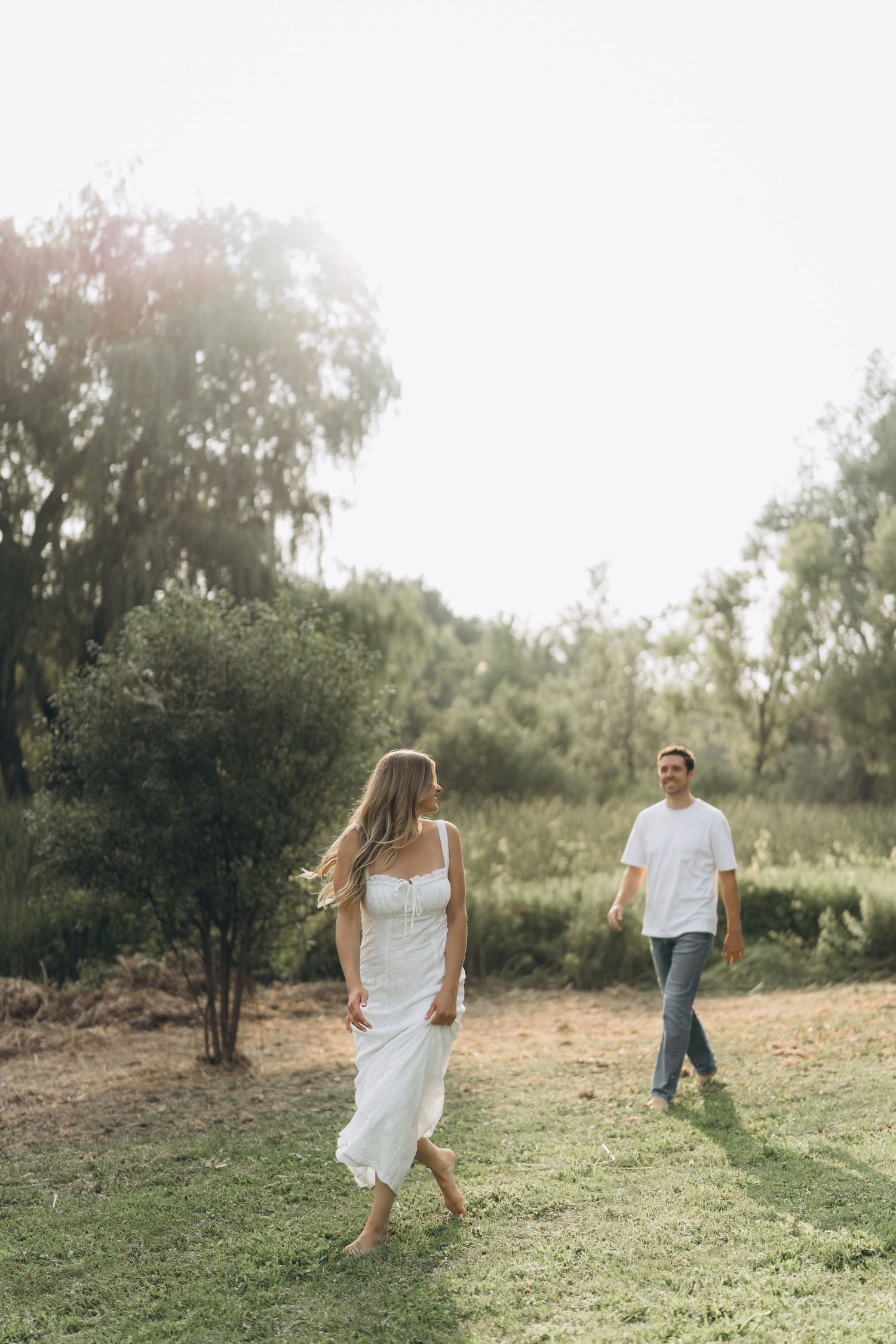 A couple in white outfits playfully runs through a grassy field on a sunny summer day.