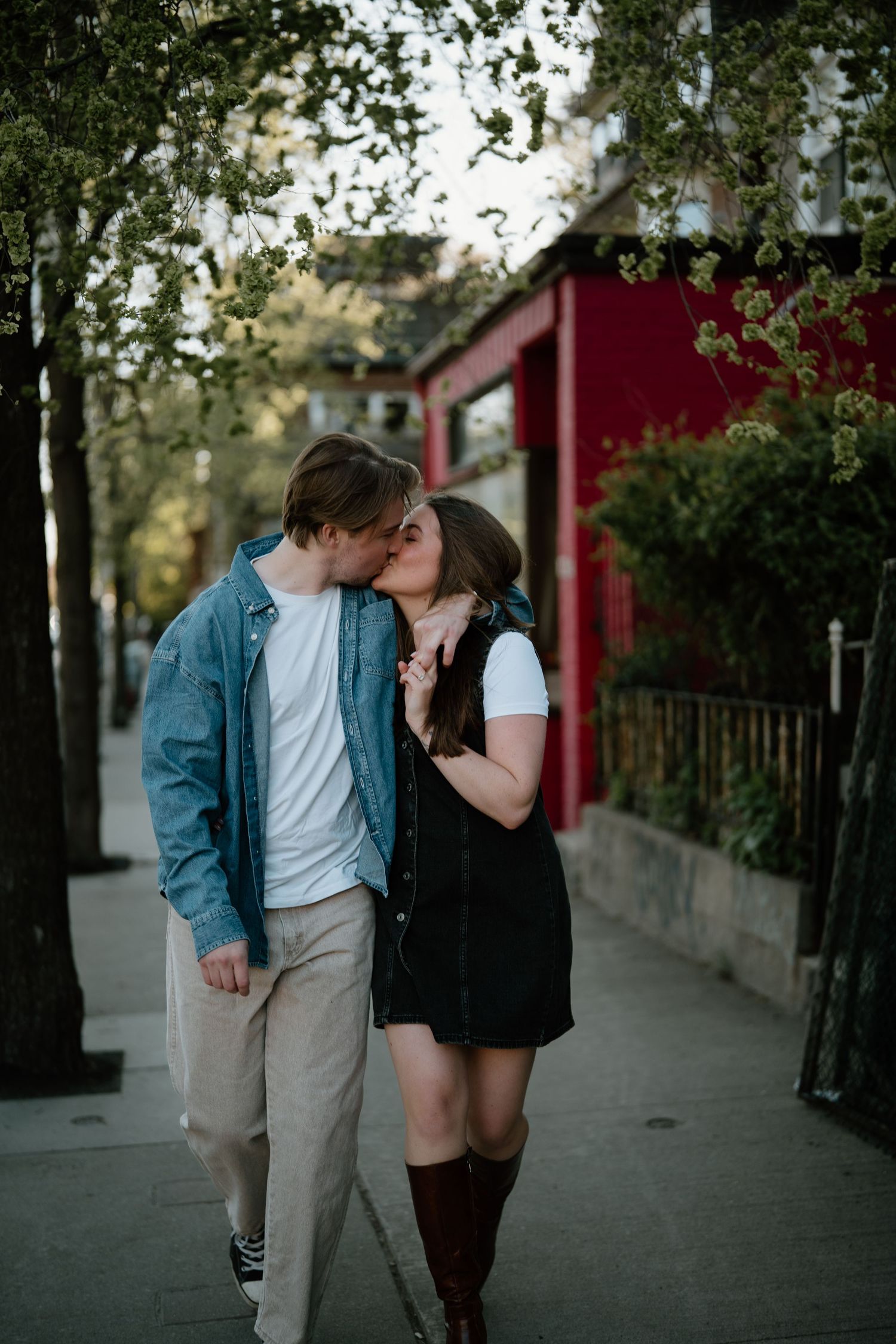 A romantic moment shared between a couple on a sidewalk near a red building on a warm evening.