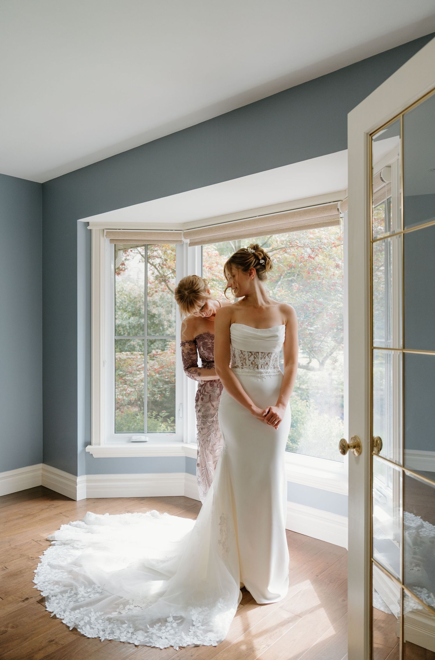 Two people in elegant white gowns share an intimate moment by a sunlit window in a sage green room.
