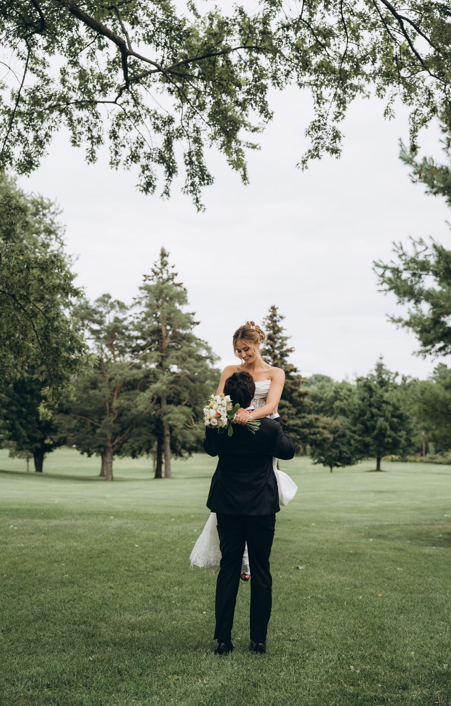 A couple embraces on a lush green lawn surrounded by trees during an outdoor wedding photoshoot.