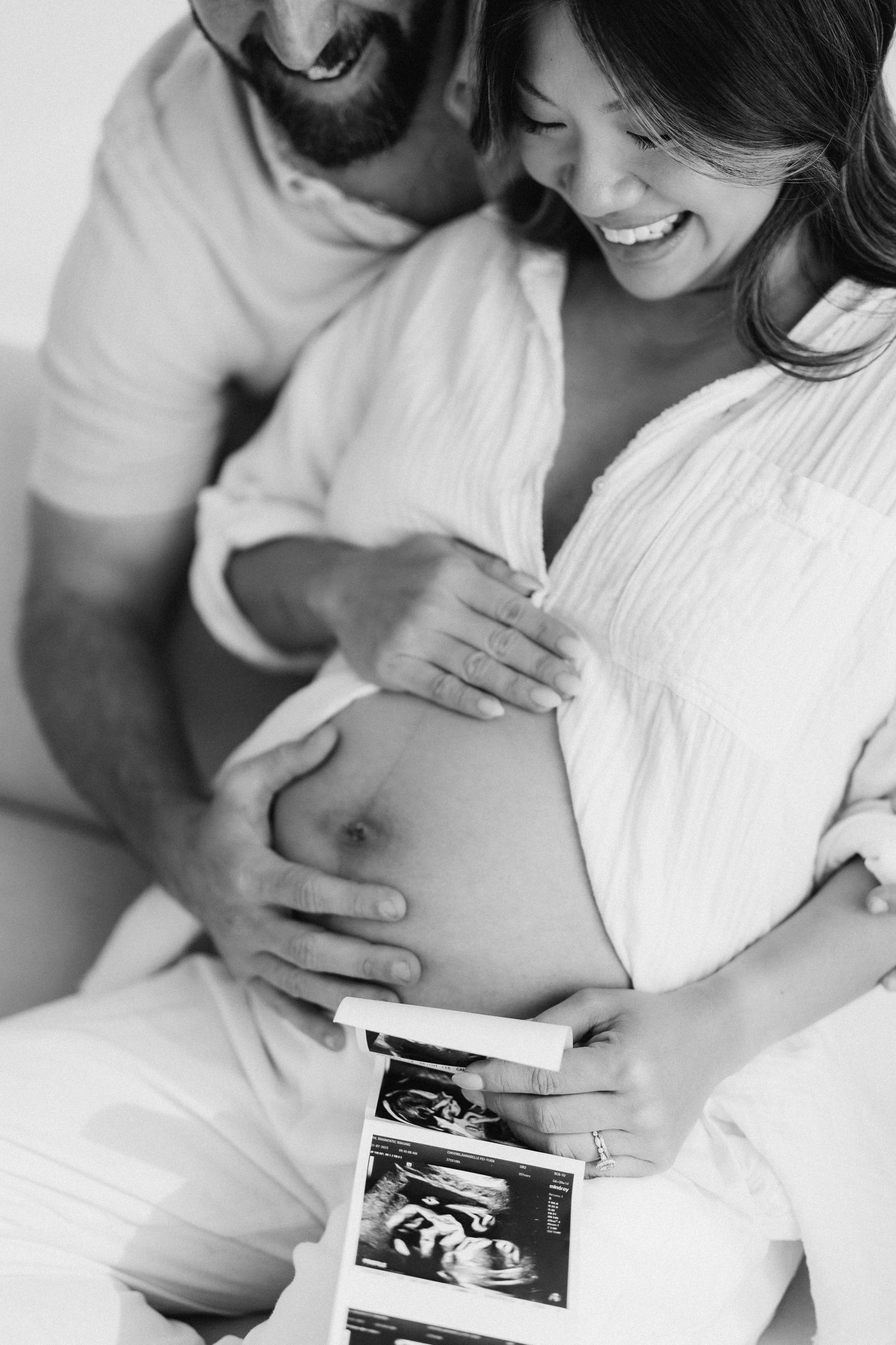 Black and white photo of expecting couple looking at ultrasound photo while touching pregnant belly in white clothing.