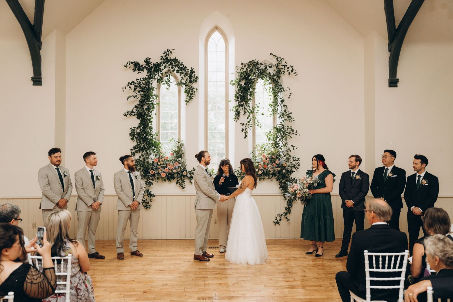 Wedding ceremony guests stand in a bright white chapel with greenery decorations and tall windows.