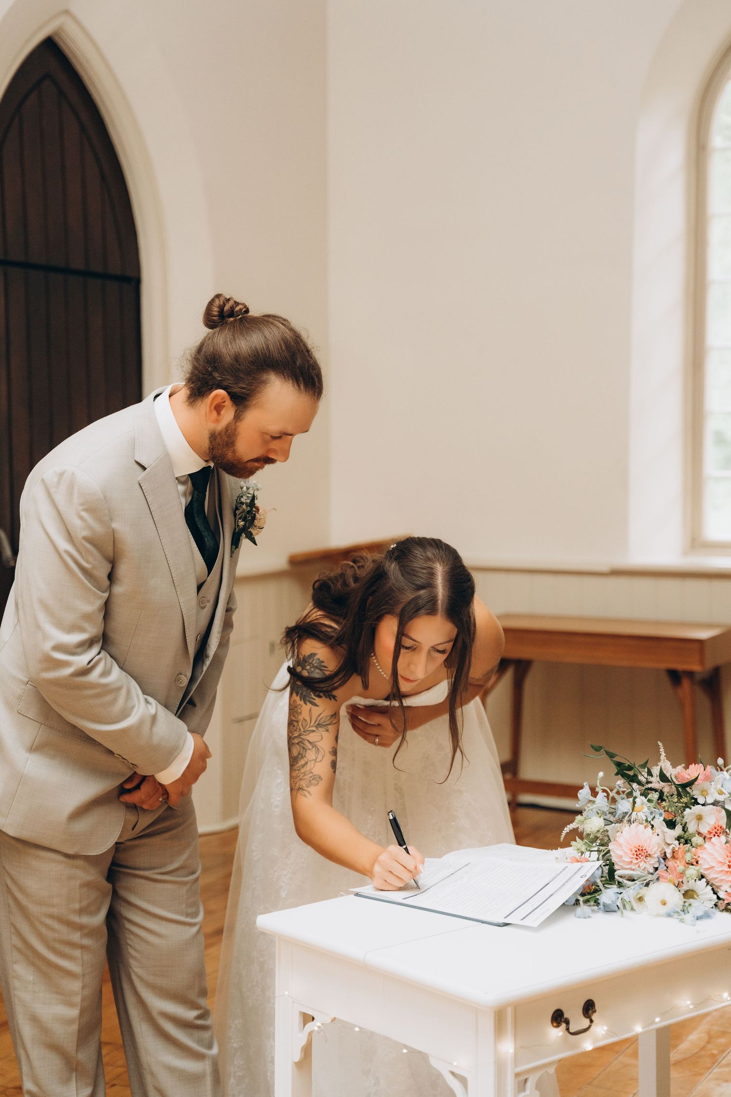 A couple signs their marriage documents at a white altar table in a church setting with pink floral arrangements.