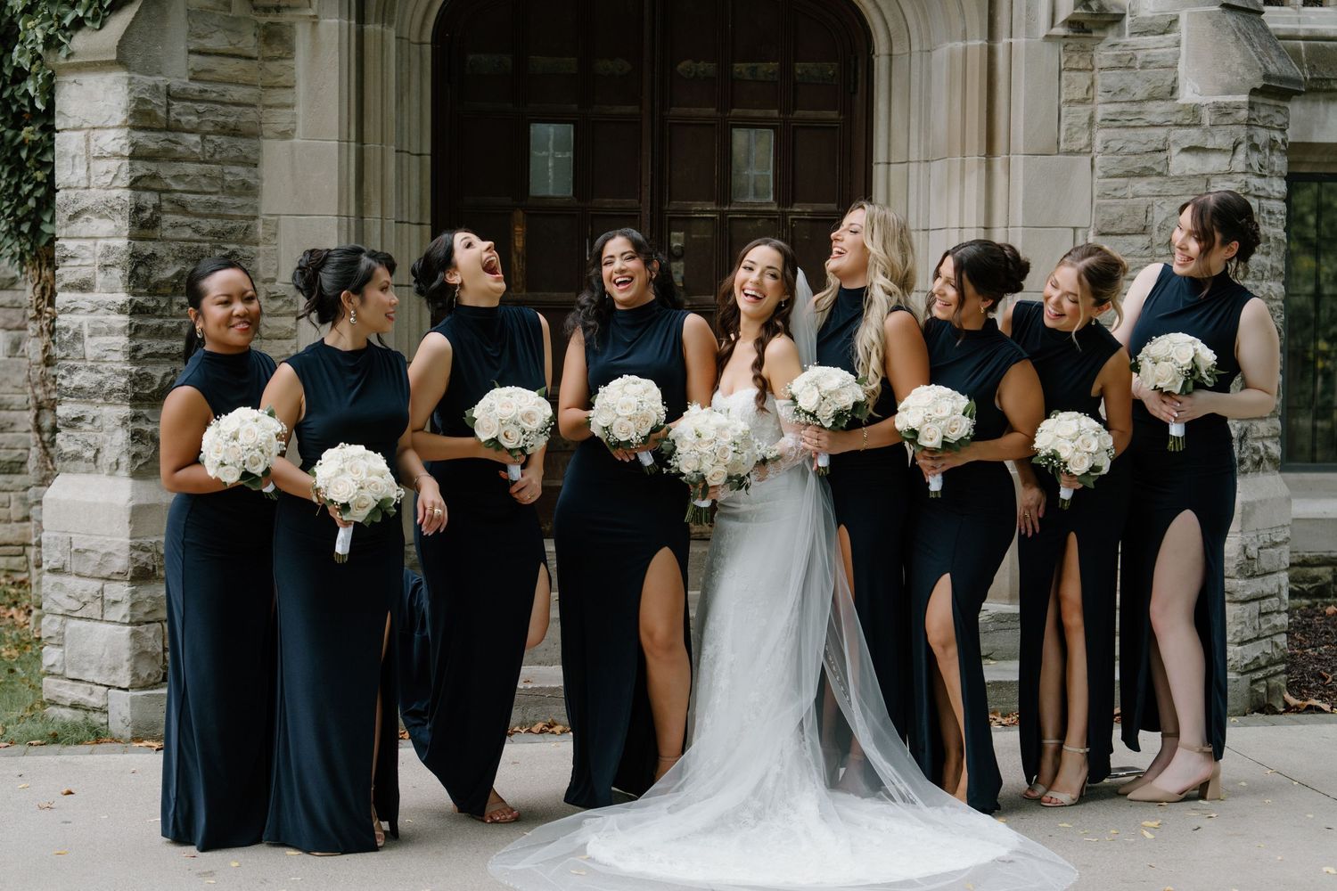 Wedding party in navy blue bridesmaid dresses and white wedding gown pose together holding white bouquets outside stone church.