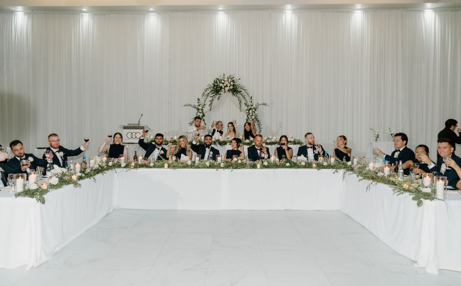 A panoramic view of an elegant wedding reception venue with white drapes, spotlights and long dining tables.