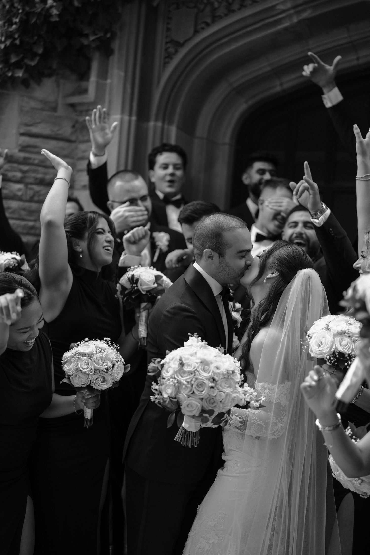 A black and white wedding photo showing a joyous celebration with bridesmaids holding white rose bouquets as the couple kisses.