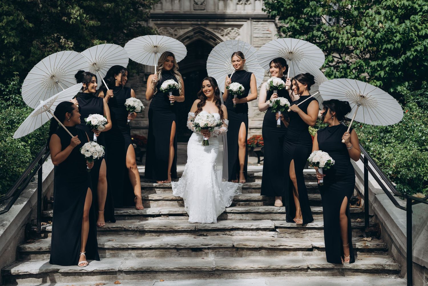 Wedding party in black dresses and white parasols pose elegantly on stone steps during outdoor ceremony.
