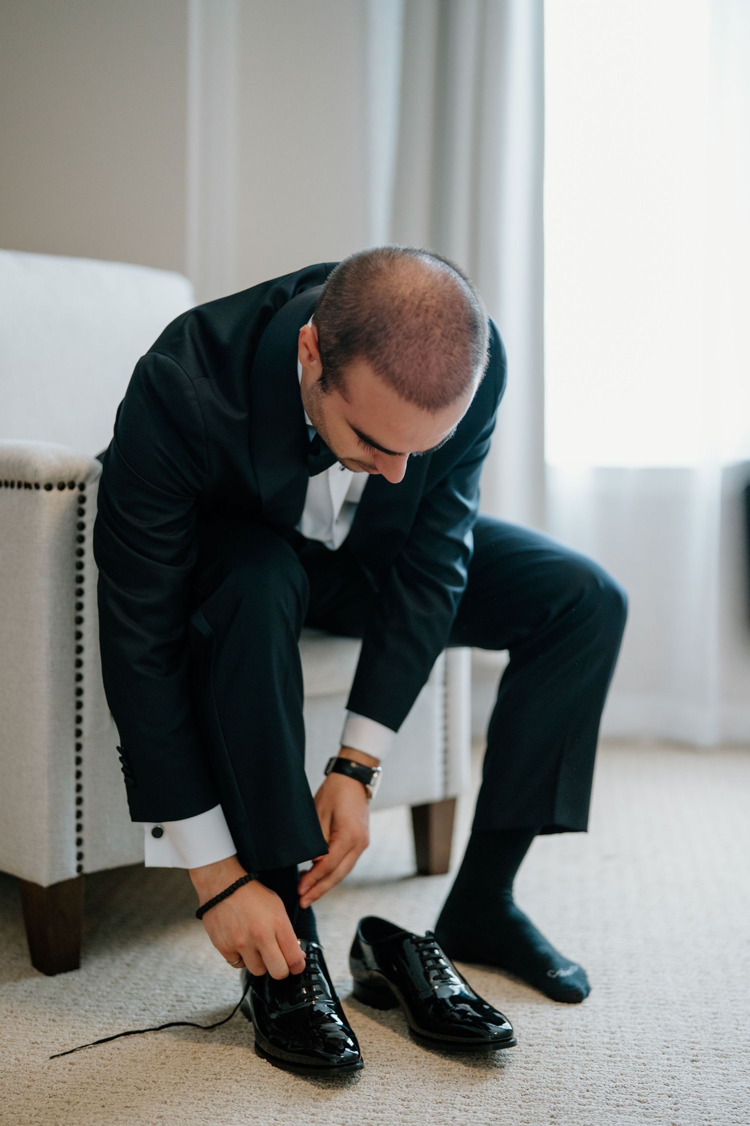 Person in dark suit sits on upholstered chair while tying formal black dress shoes on light carpet.