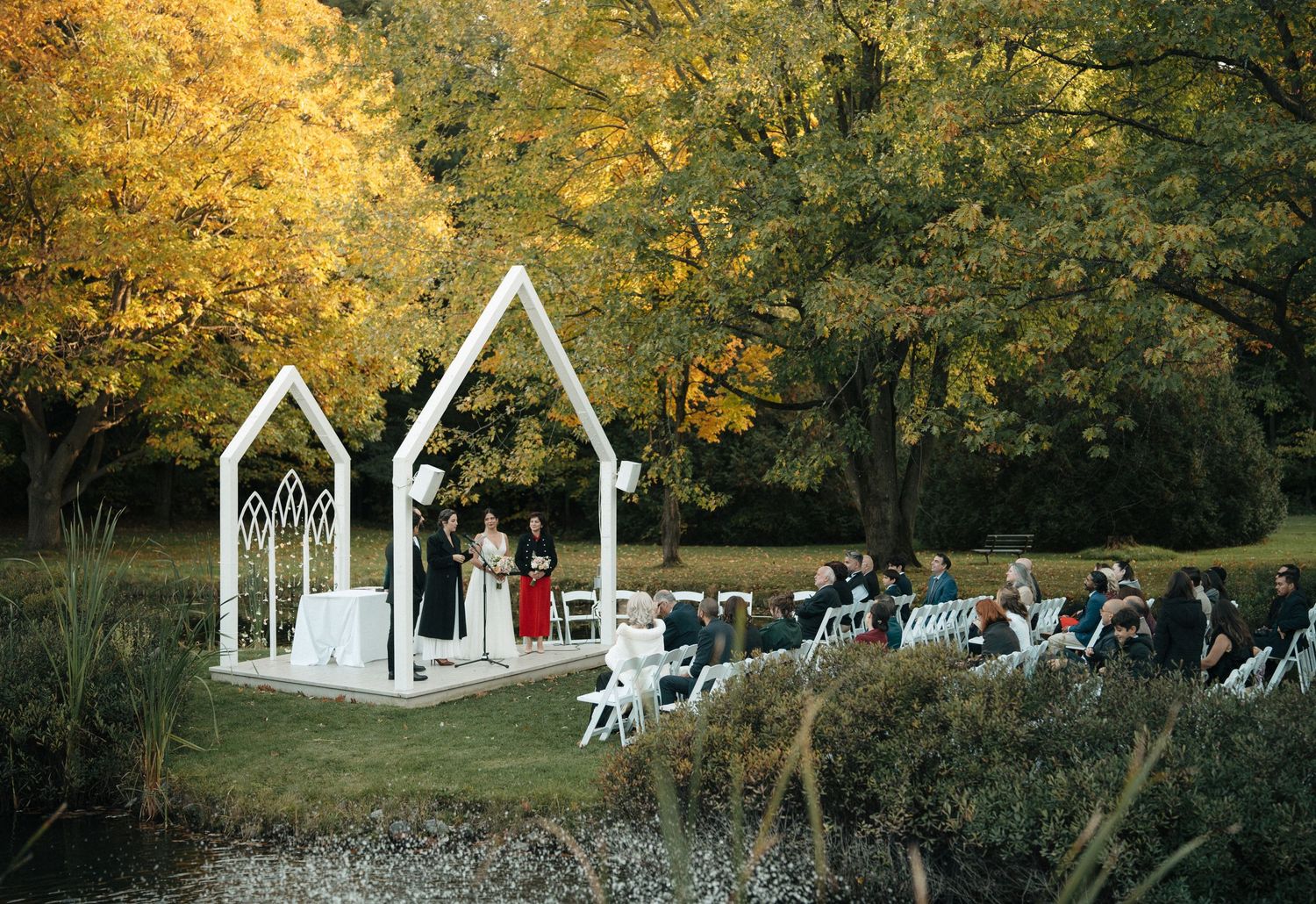 Wedding ceremony in progress under white garden arches with autumn foliage backdrop.