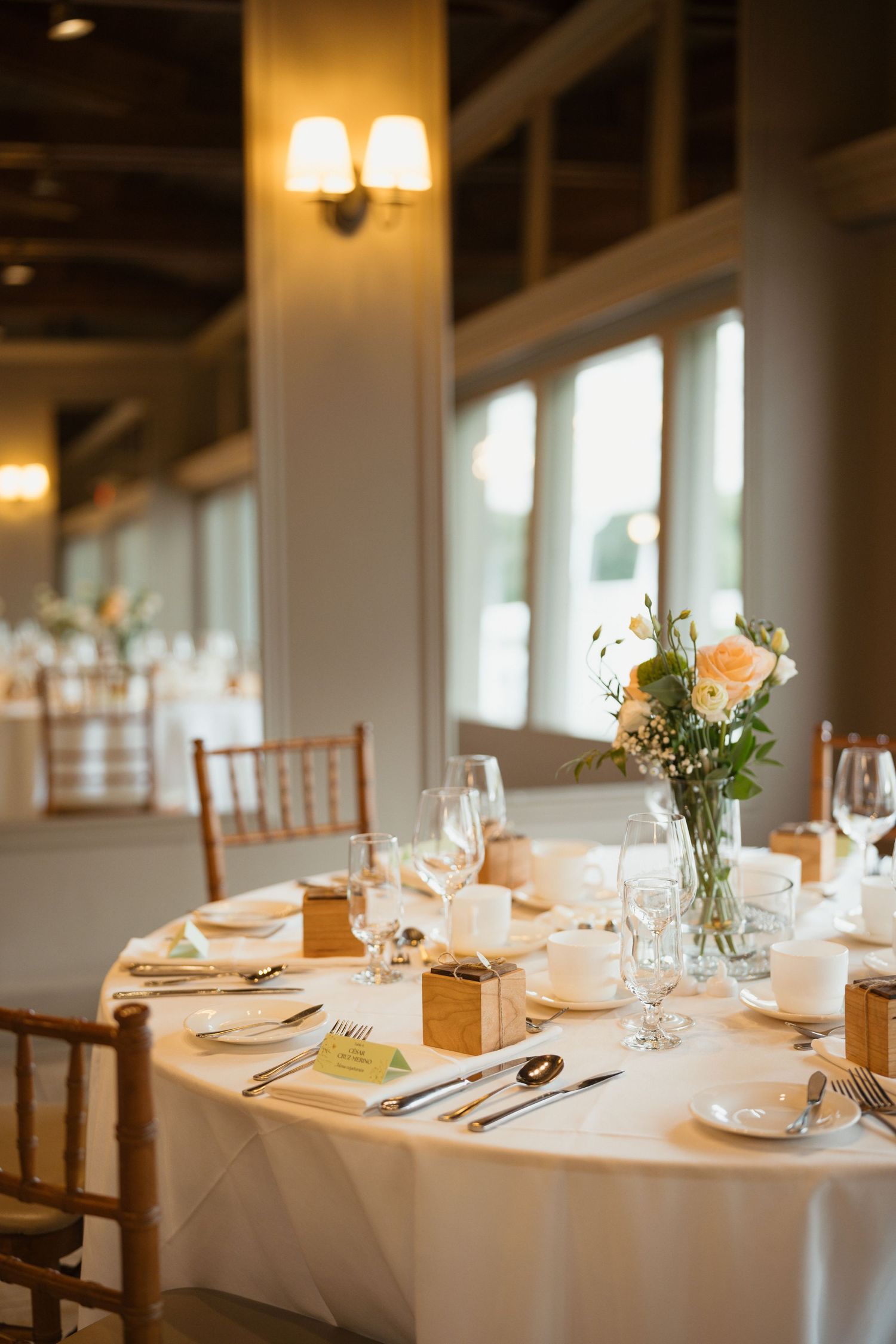 Sunlit wedding reception table with wooden Chiavari chairs, white linens, and peach floral centerpiece.