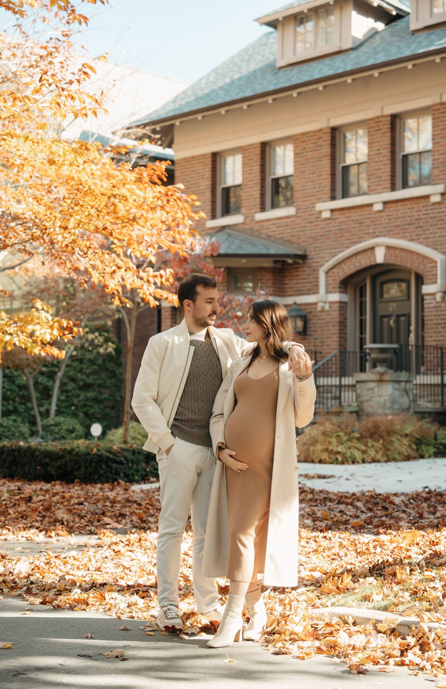 Couple strolls along brick building surrounded by fallen autumn leaves in warm afternoon light.