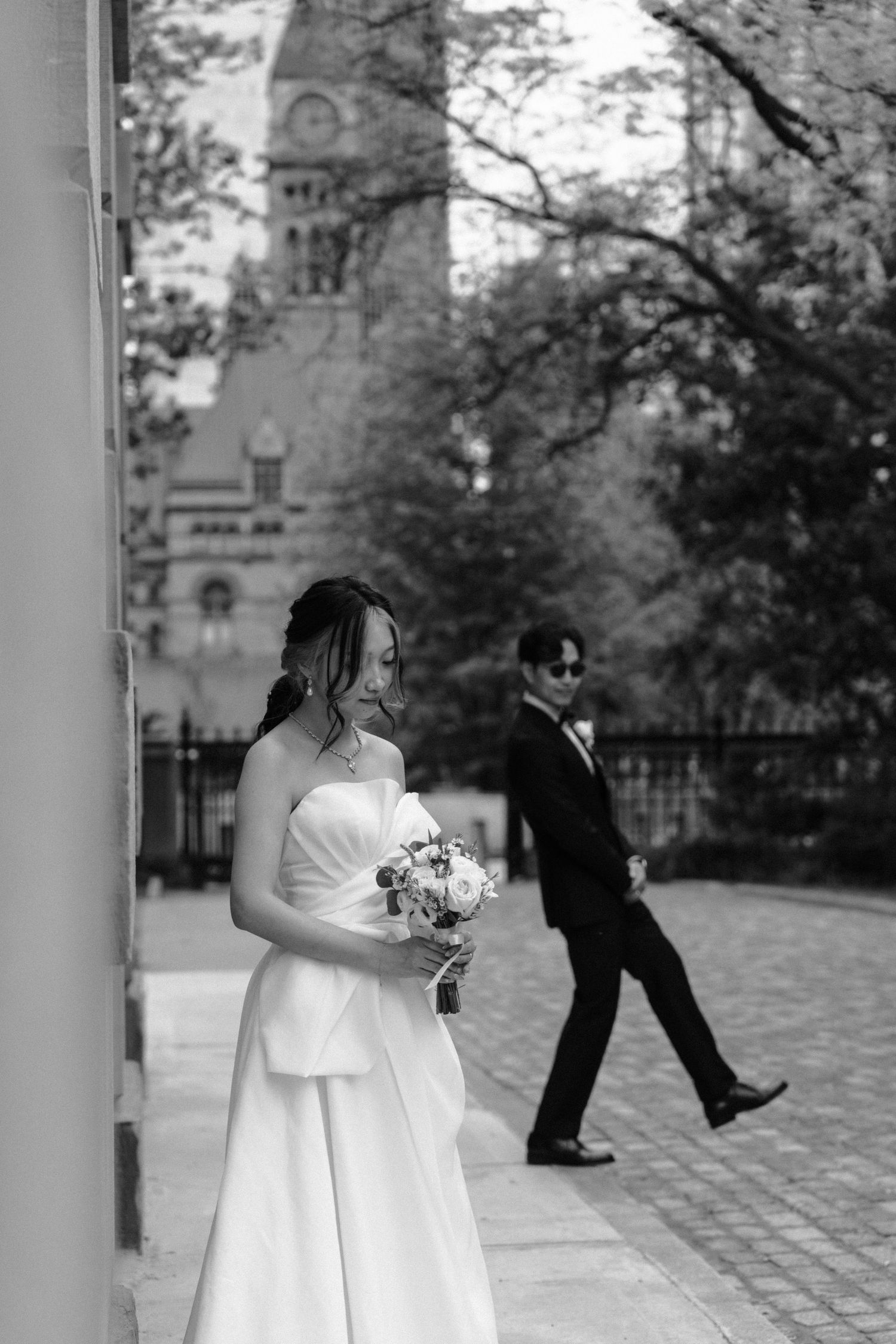 Black and white wedding photo with bride holding bouquet while groom strides across courtyard near church tower.