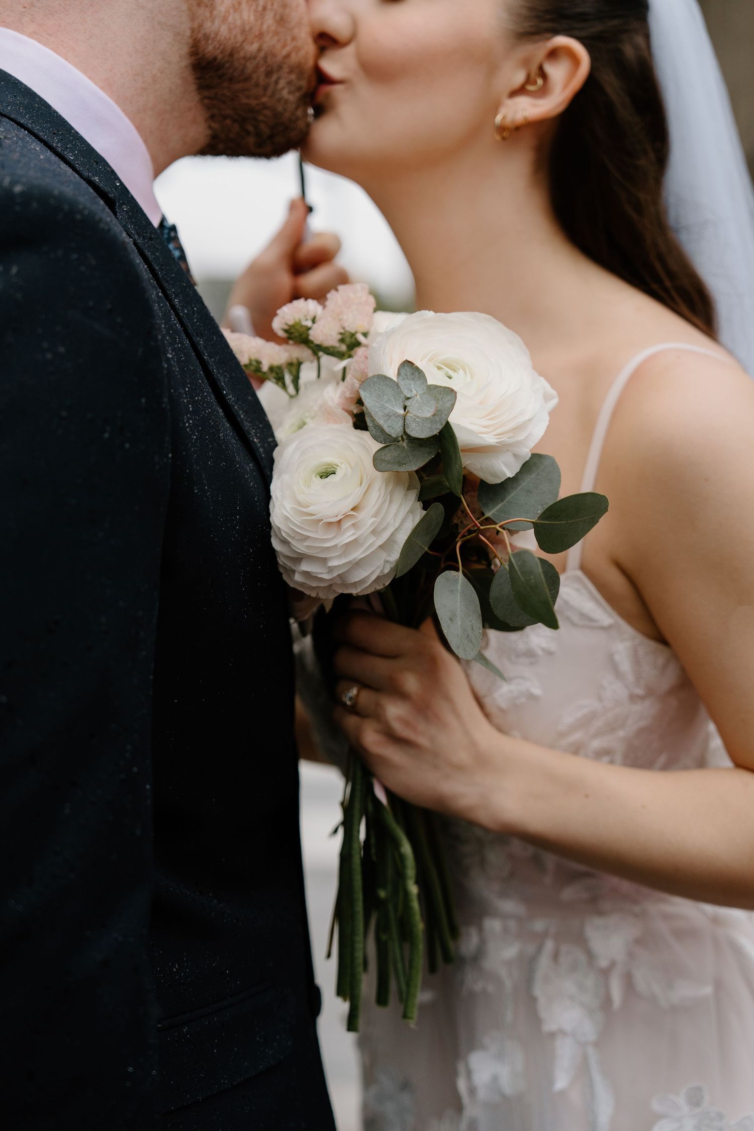 Close up of bride holding a delicate white ranunculus and eucalyptus bouquet while kissing during an intimate wedding moment.