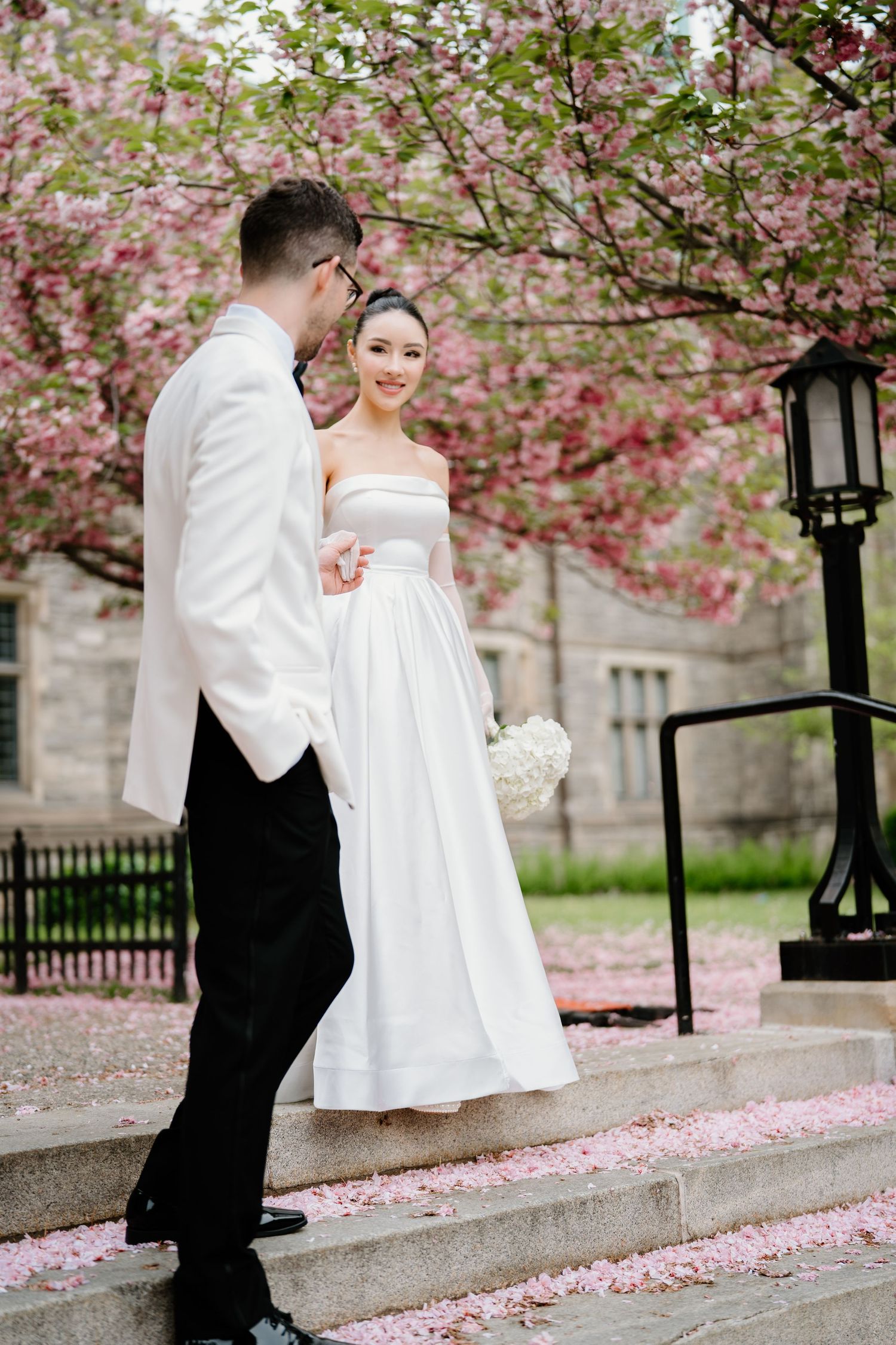A couple in formal wedding attire pose on stone steps surrounded by blooming pink cherry blossoms and a vintage lamp post.