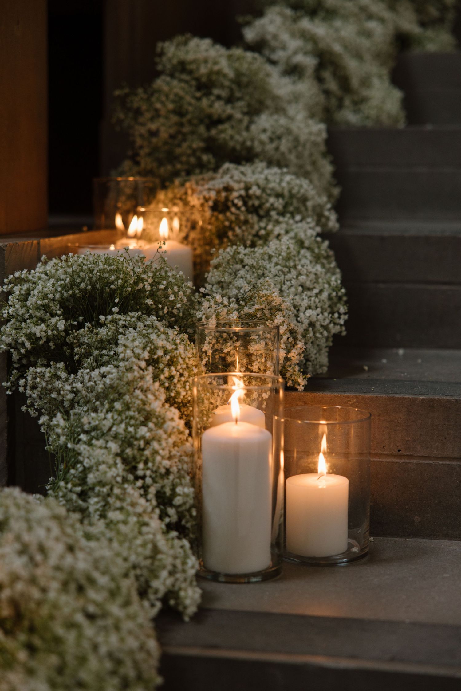 Glowing white candles in glass holders line wooden stairs decorated with delicate baby's breath flowers and greenery.